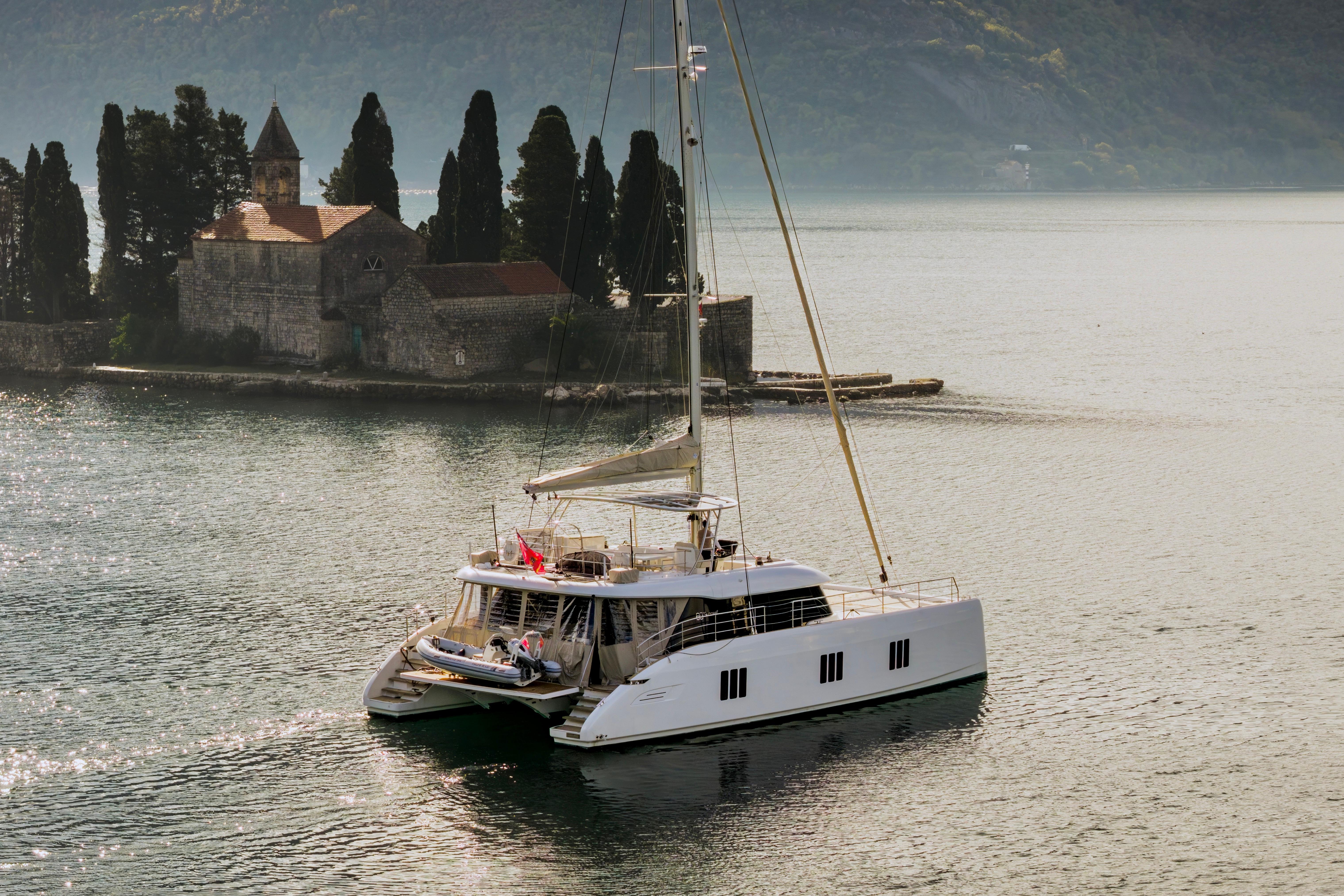 2023 Sunreef 60 catamaran sailing near a historic stone building and cypress trees.