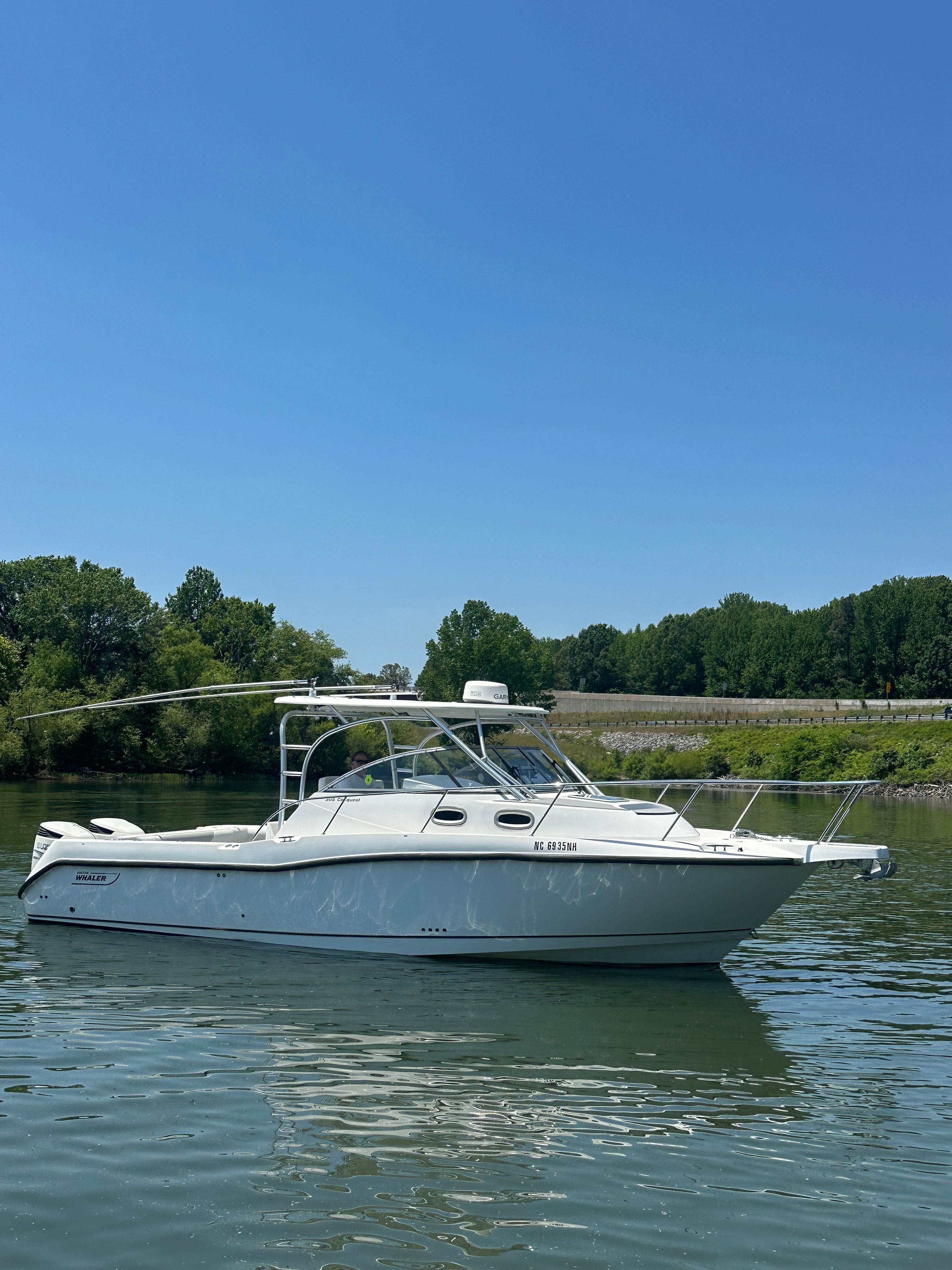 A white motorboat on calm water, surrounded by green trees under a clear sky.