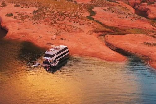 Houseboat "The Queen Elizabeth" by Adonia, 2021, on serene water near red rocky shore.