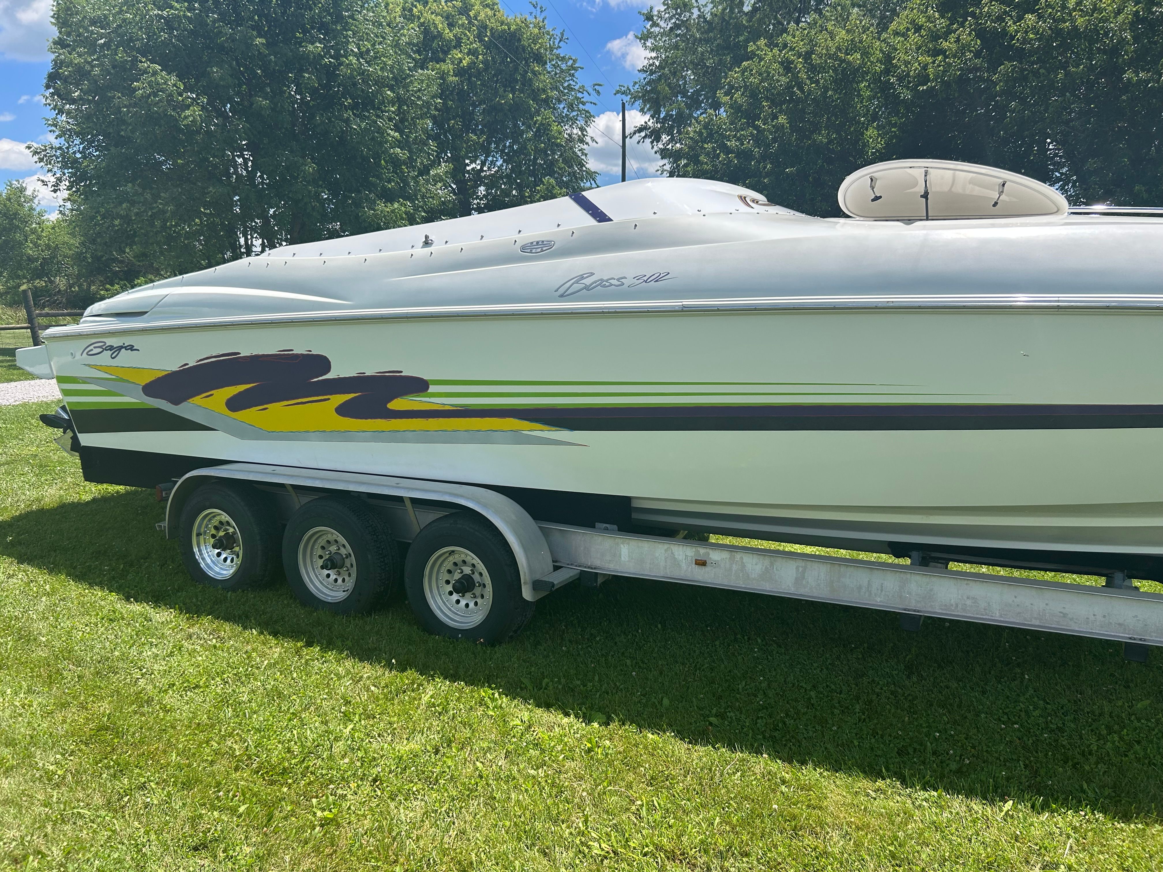A 1996 Baja 302 boat on a trailer, surrounded by grass and trees.