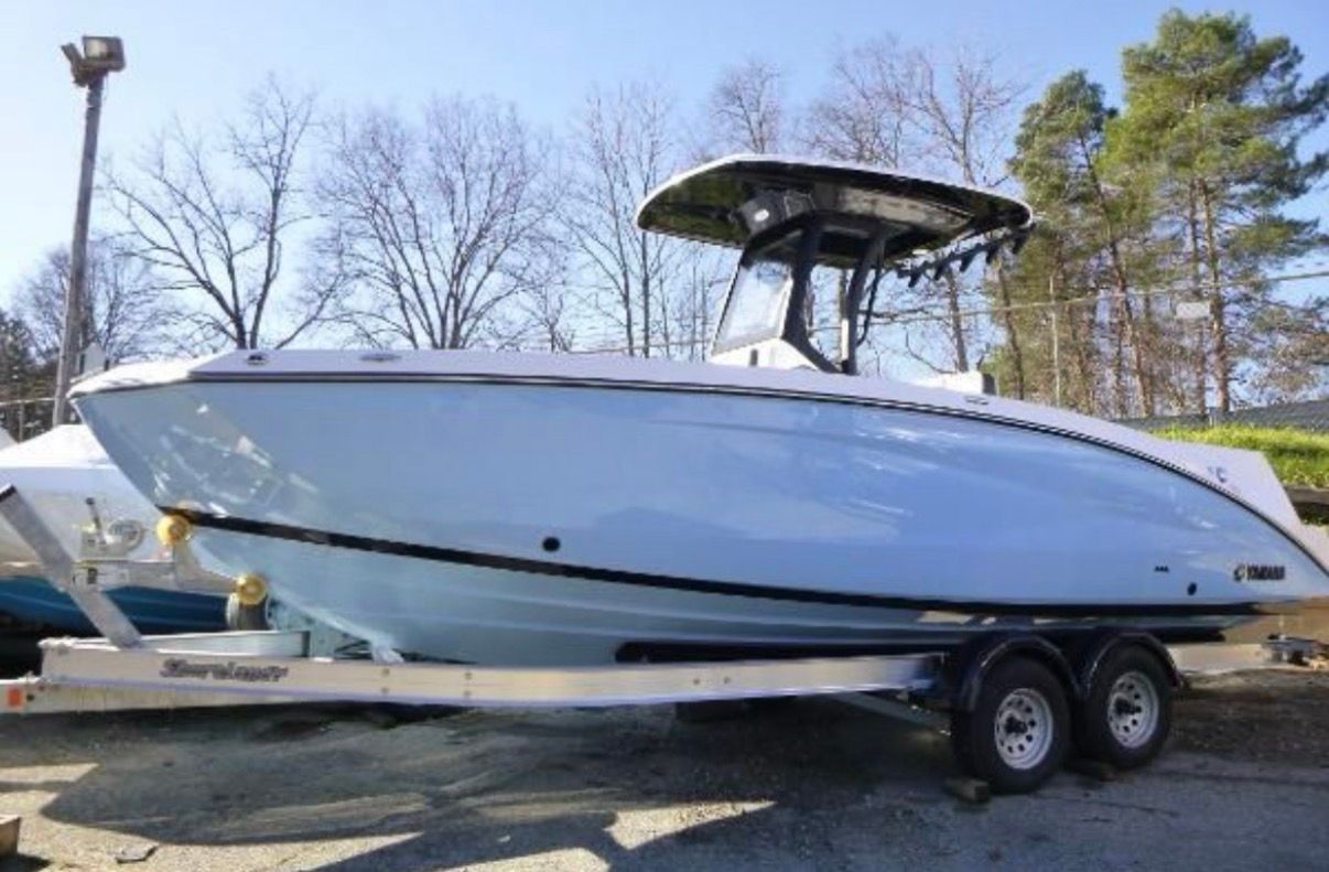A 2024 Yamaha 255 FSH Sport H boat on a trailer, surrounded by trees and clear sky.