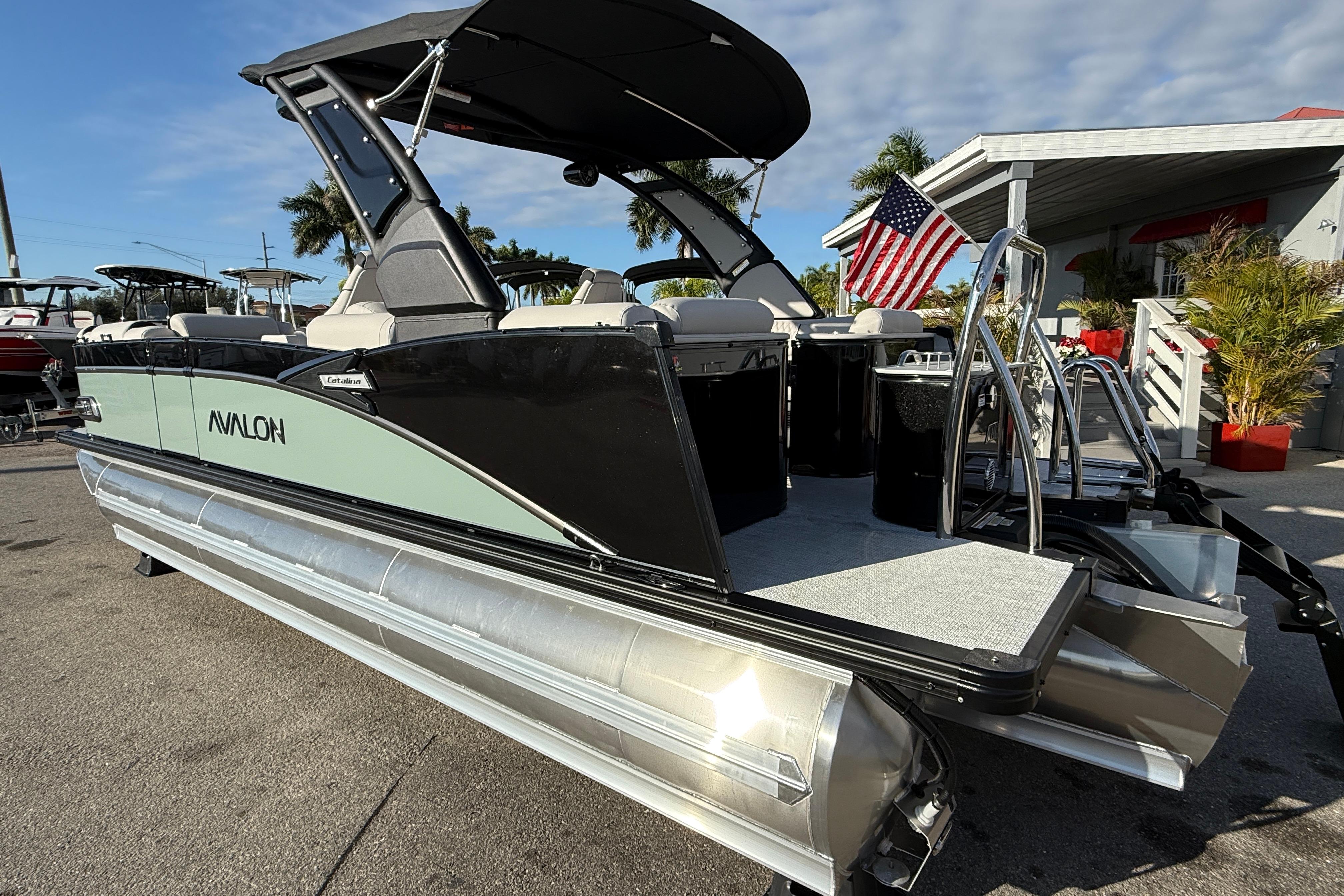 2026 Avalon 2385 Catalina Quad Lounger Tri Toon boat with American flag, parked outdoors.