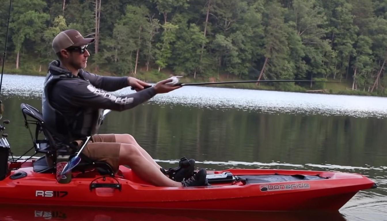 Man fishing on a red 2025 Bonafide RS117 kayak in a serene lake setting.