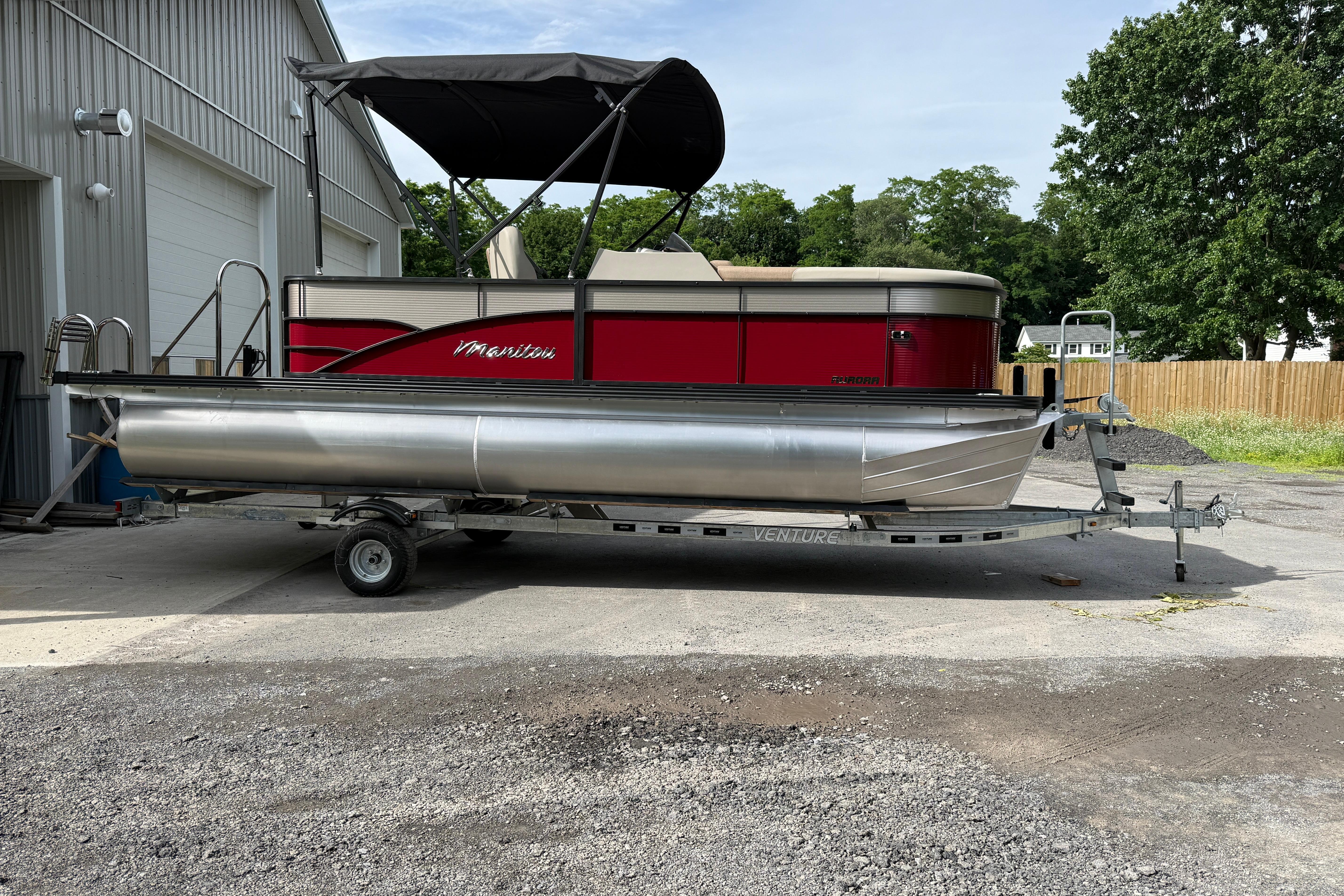 2027 Manitou 21 Aurora LE pontoon boat on trailer, parked outdoors near a building.