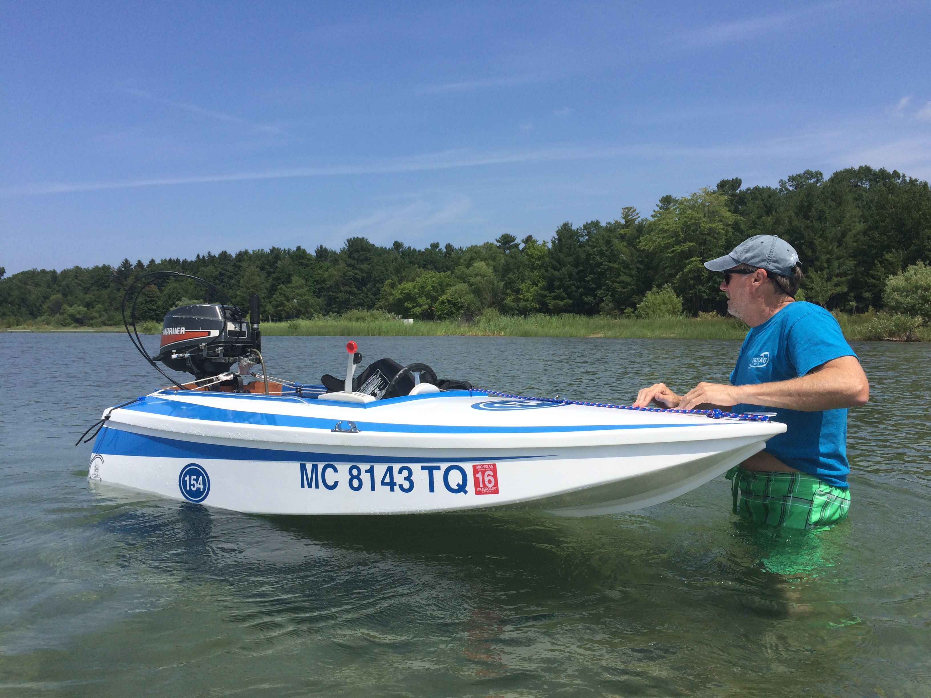 A man stands beside a small white racing boat in shallow water.