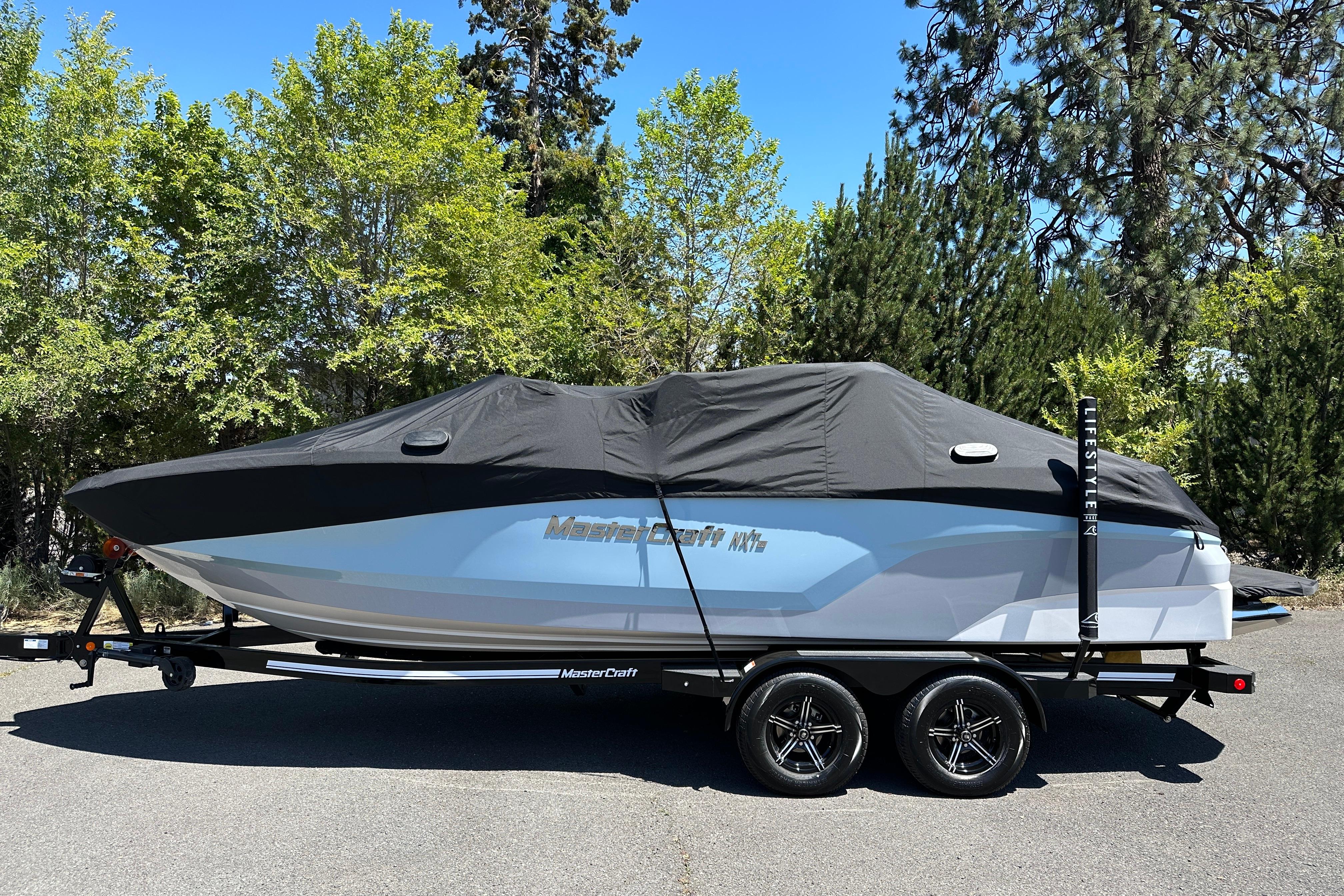 2023 MasterCraft NXT23 boat covered on trailer, parked outdoors with trees in background.