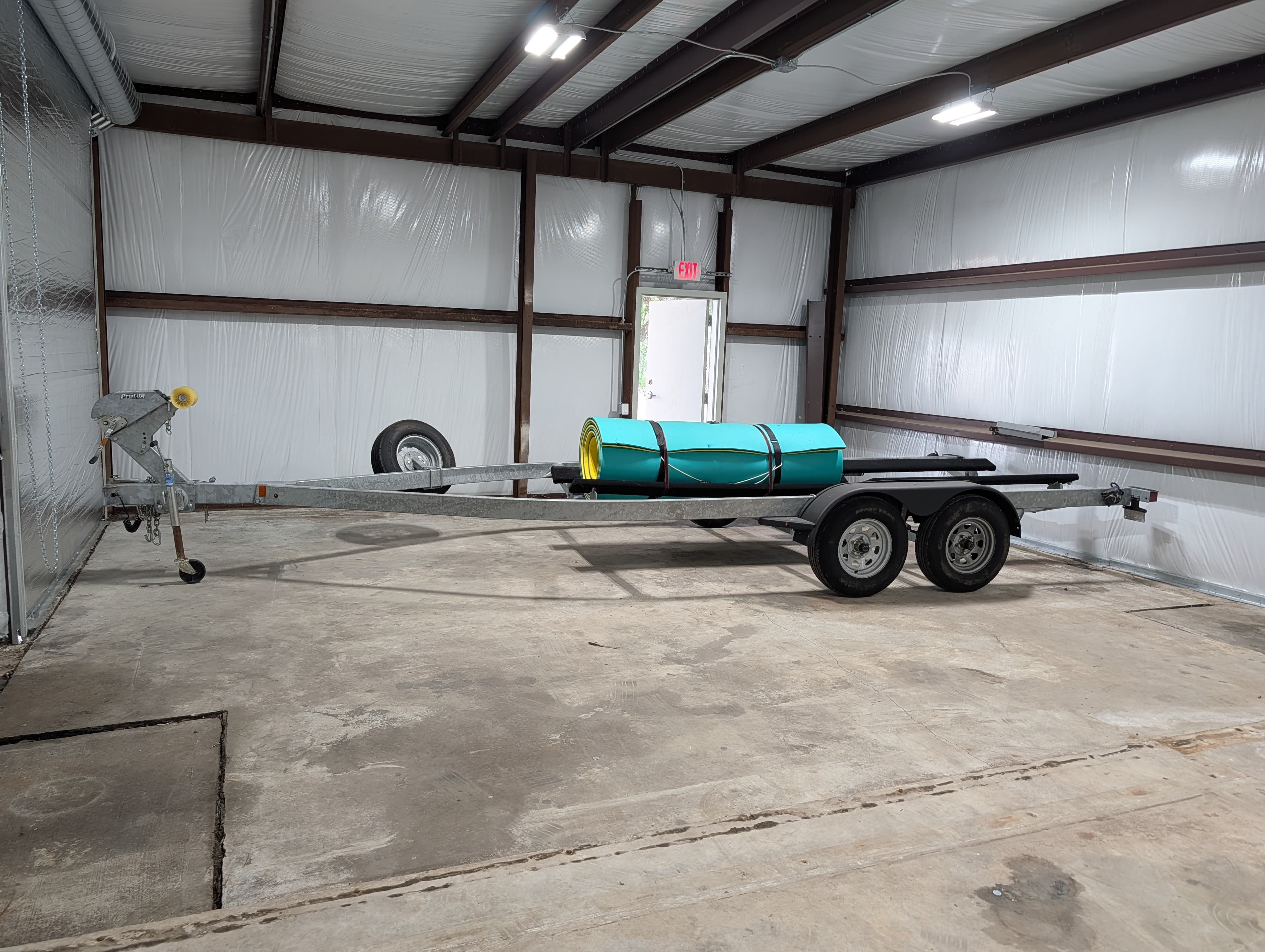 An empty boat trailer is in a warehouse. It has two axles and a spare tire.