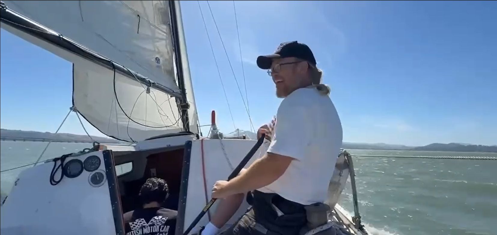 A man steers a sailboat on open water under sunny skies.