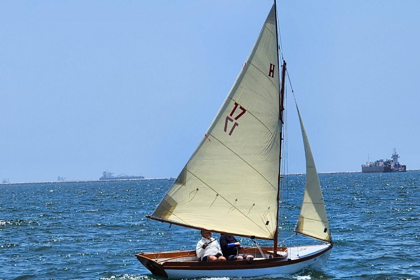 1920 Herreshoff 12.5 sailboat on open water, clear sky, distant ships visible.