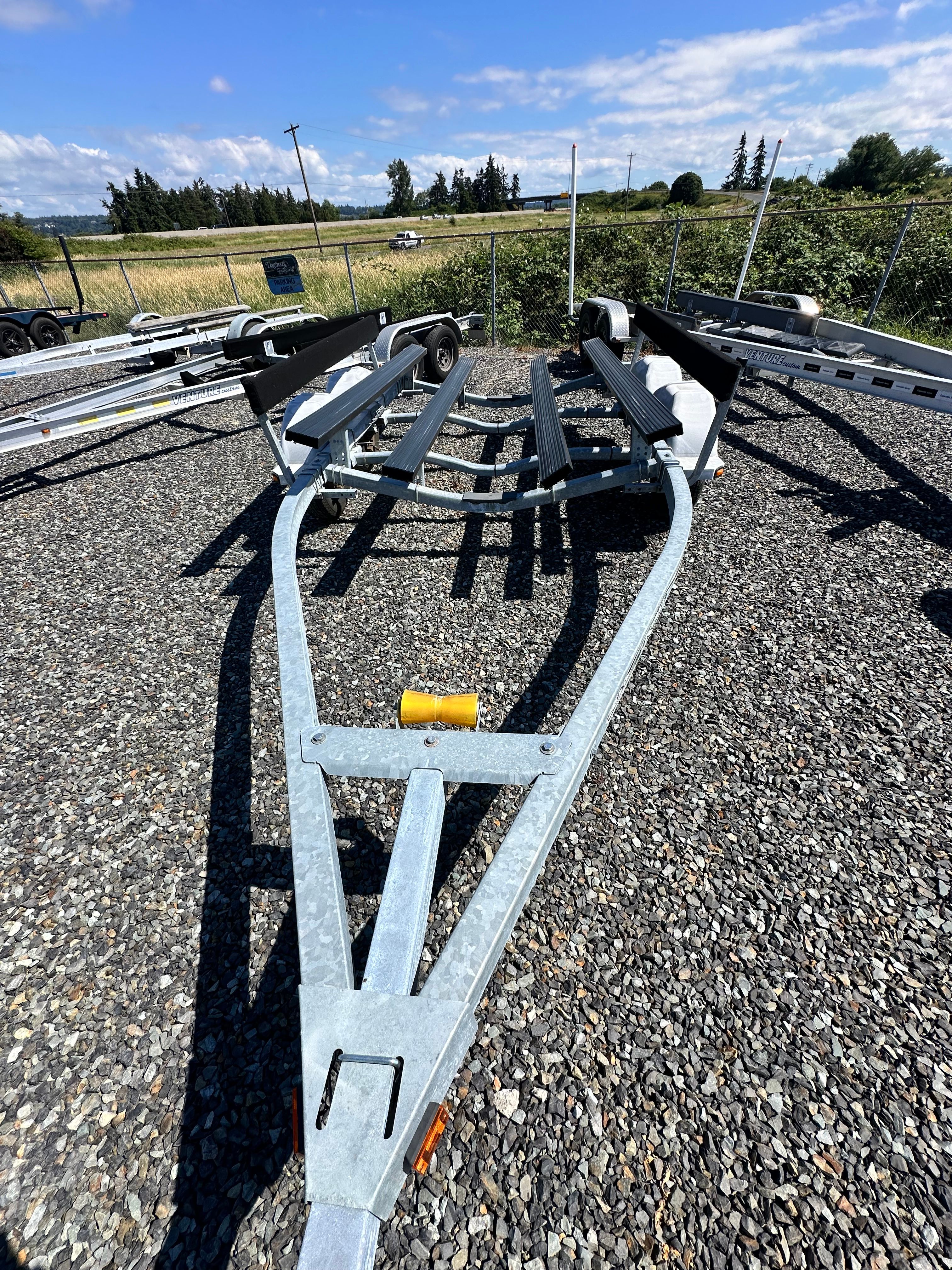 A boat trailer sits on a gravel lot, surrounded by grass and trees.