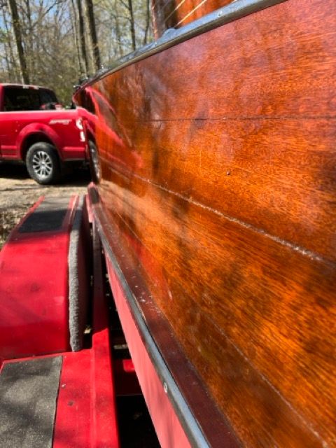 Close-up of a shiny wooden boat on a red trailer, with a red truck in the background.