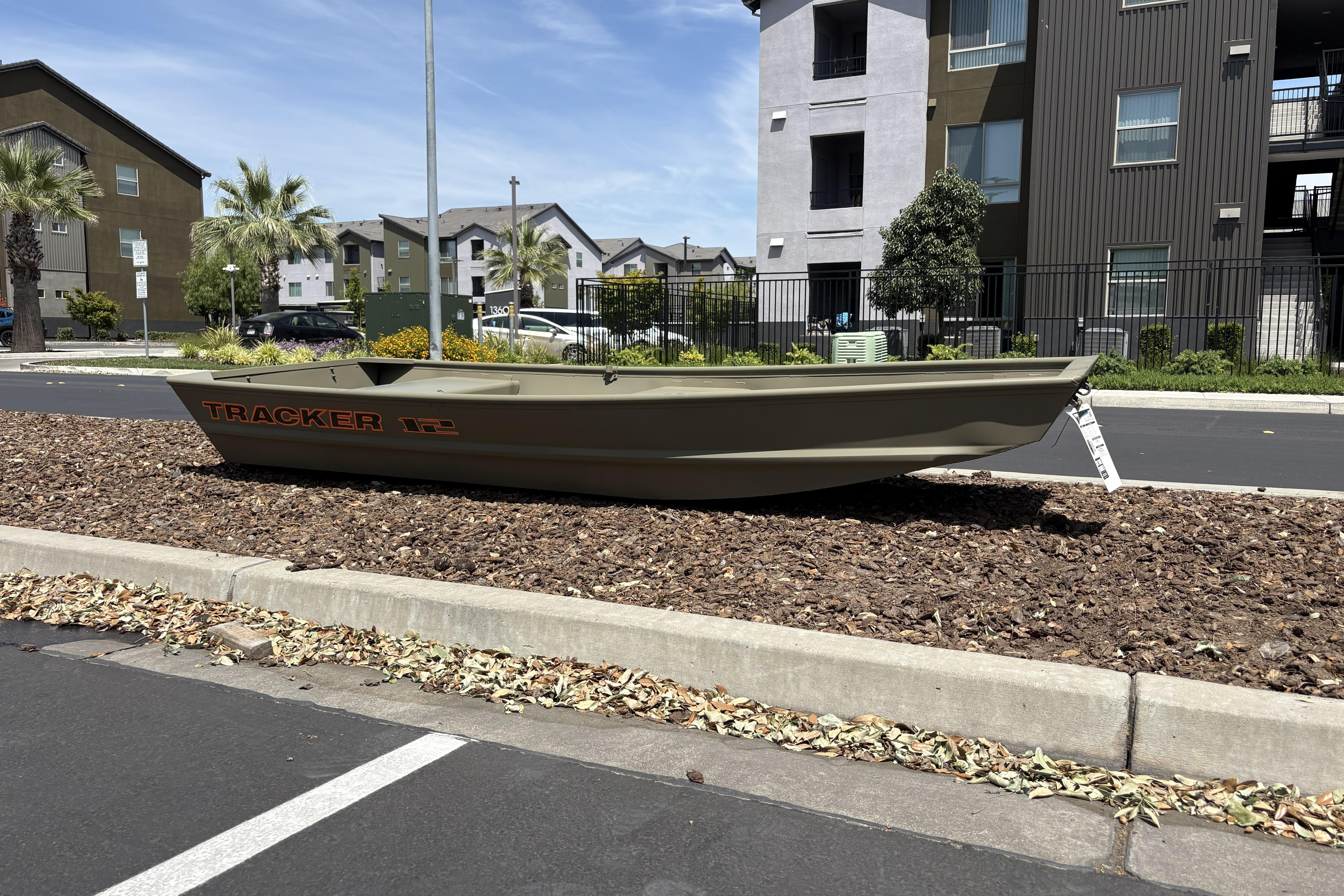 2026 Tracker Grizzly 12 Jon boat displayed on landscaped area near modern apartments.
