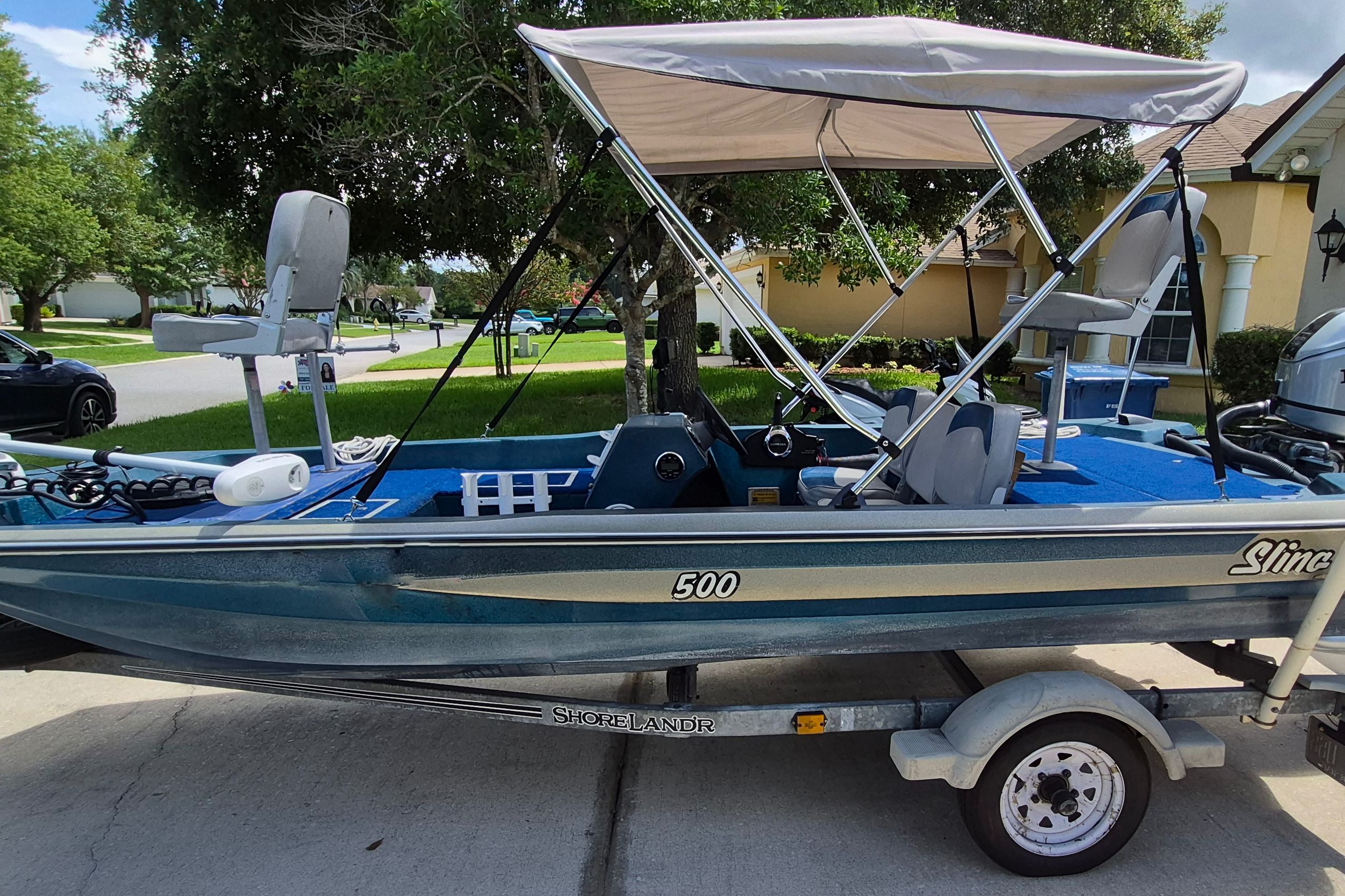 A blue boat with a sunshade is on a trailer in a residential area.