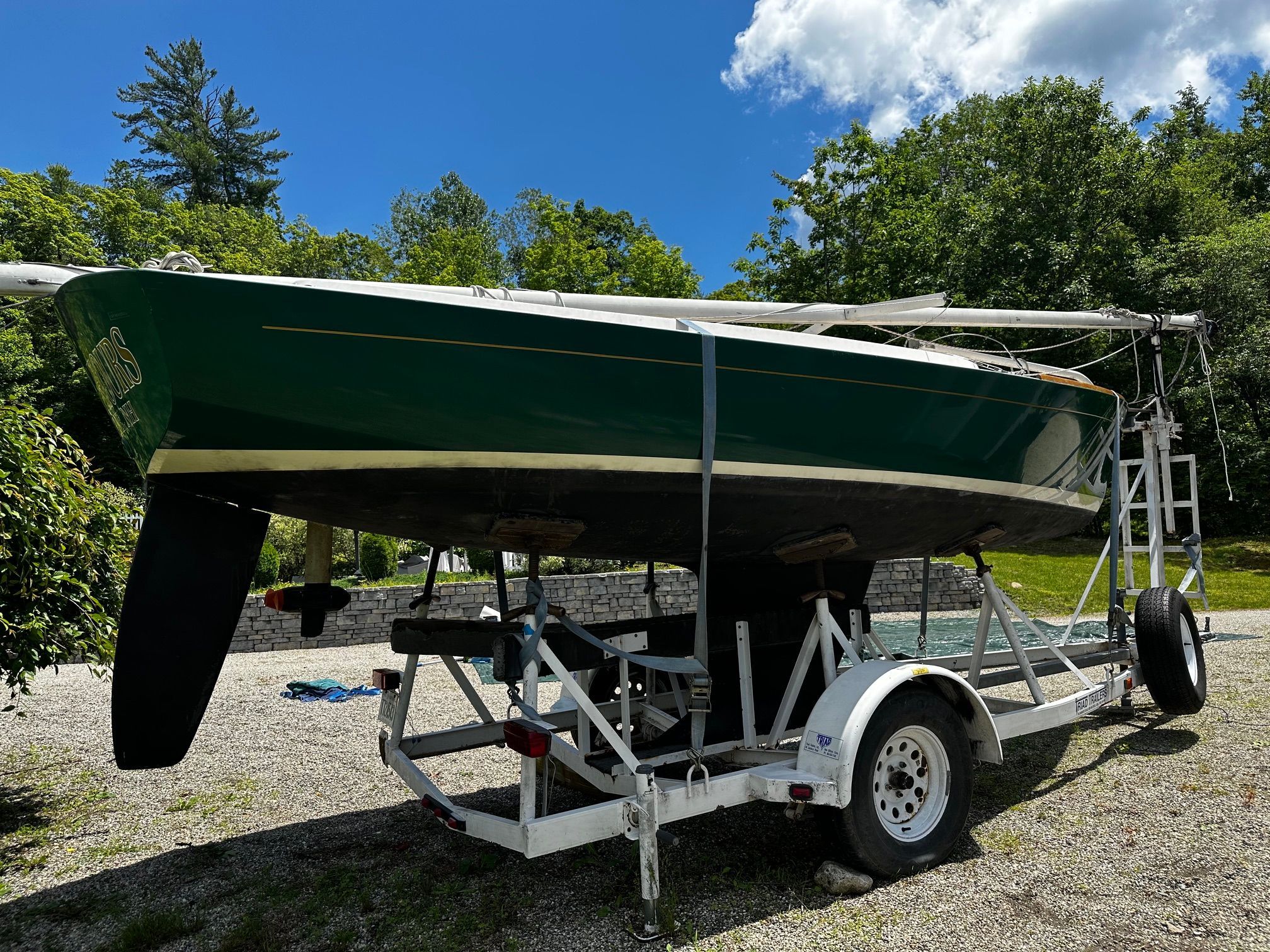 A green sailboat on a trailer, surrounded by trees and blue sky.