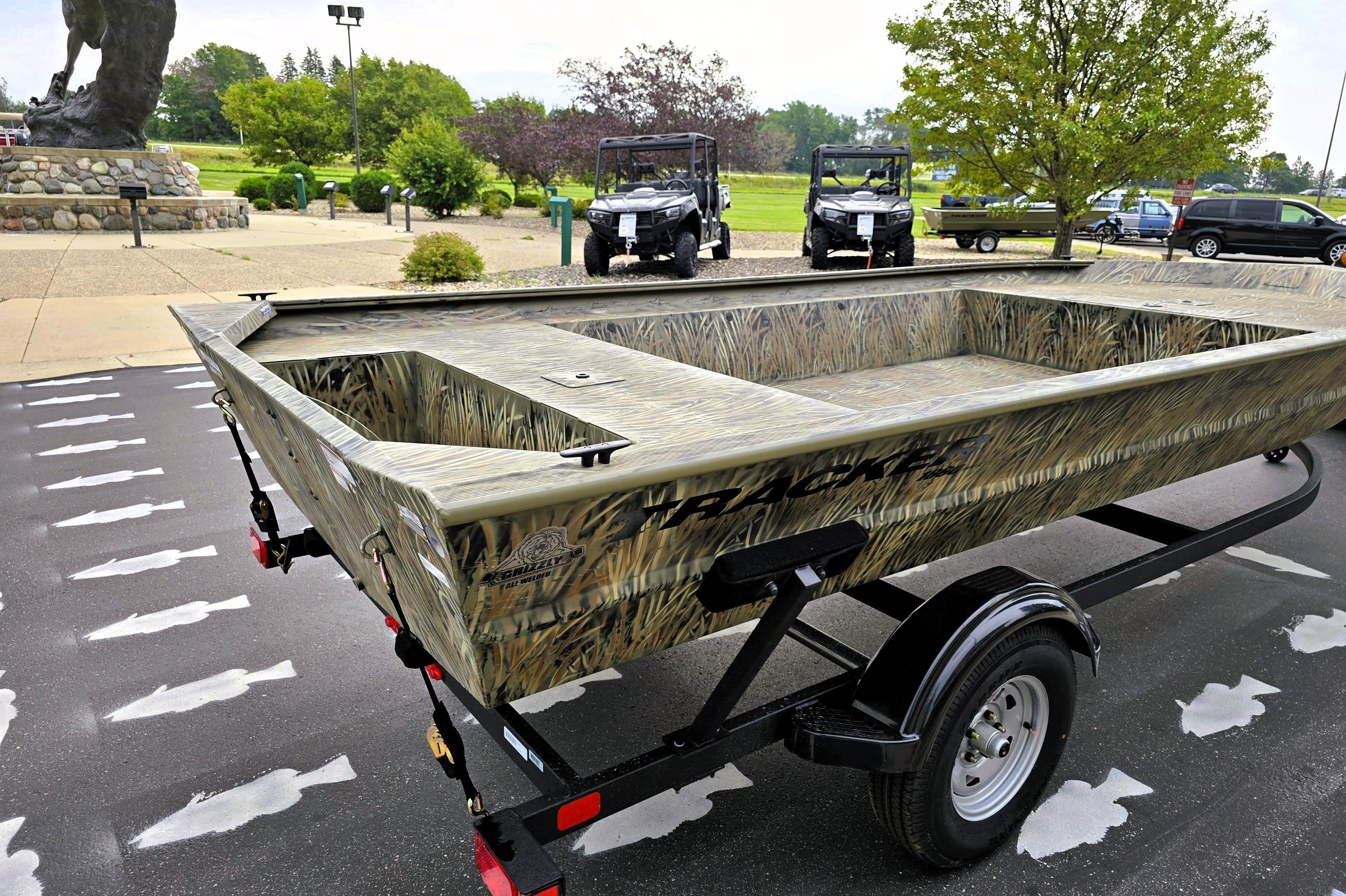 2026 Tracker GRIZZLY 1860 Utility boat on trailer, parked outdoors with trees and vehicles in background.