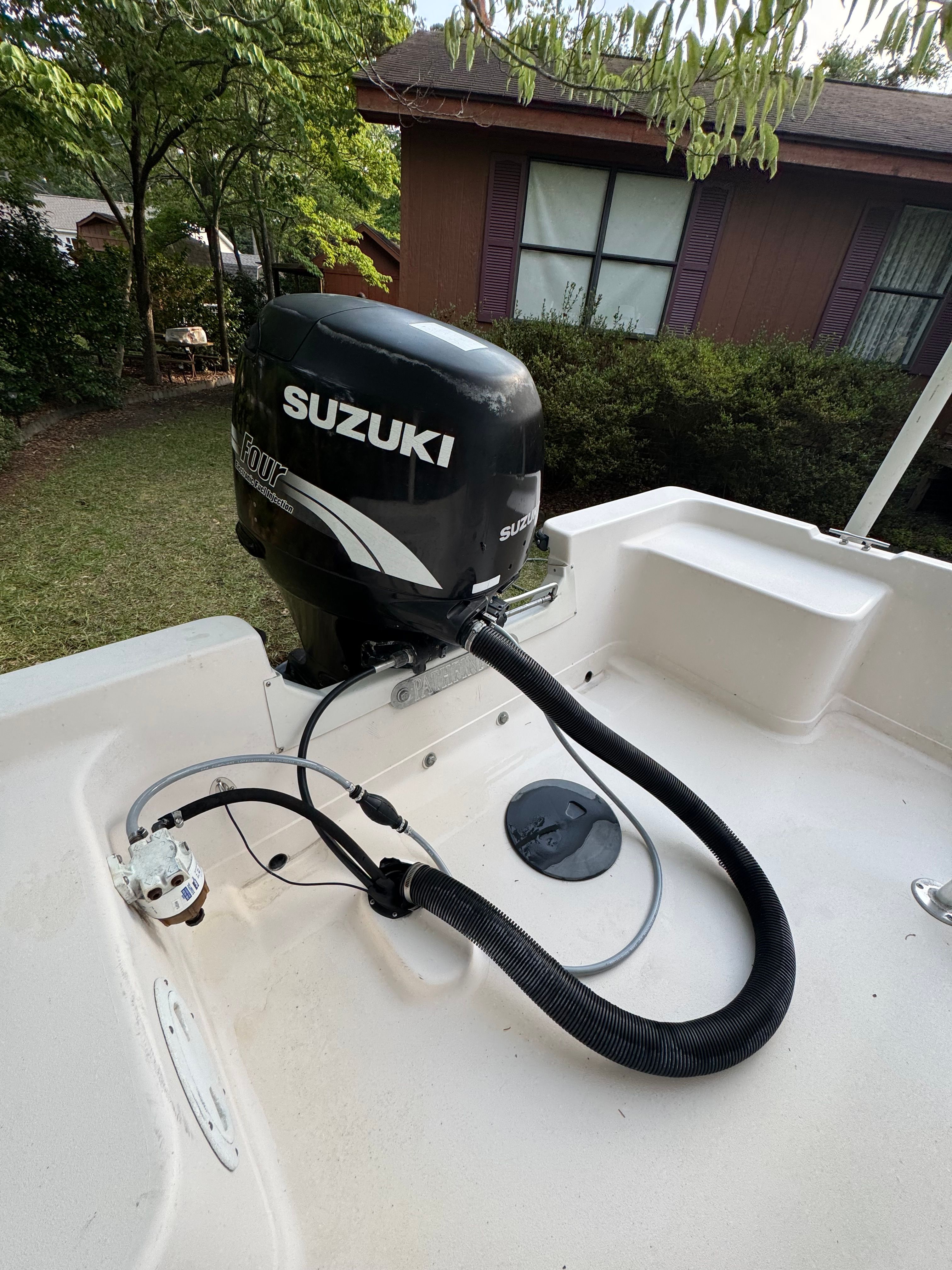 A Suzuki outboard motor is mounted on a boat, surrounded by trees and a house in the background.