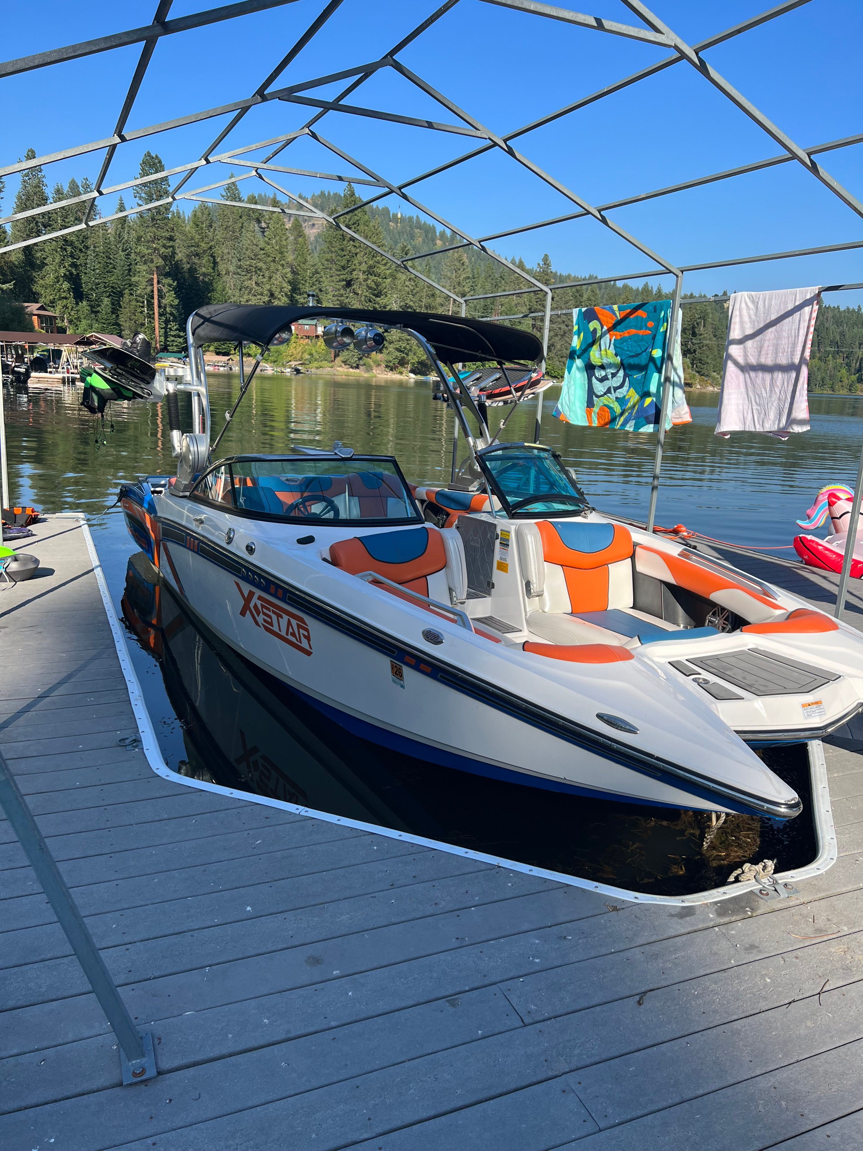 2013 MasterCraft XStar boat docked by lakeside with forest backdrop.