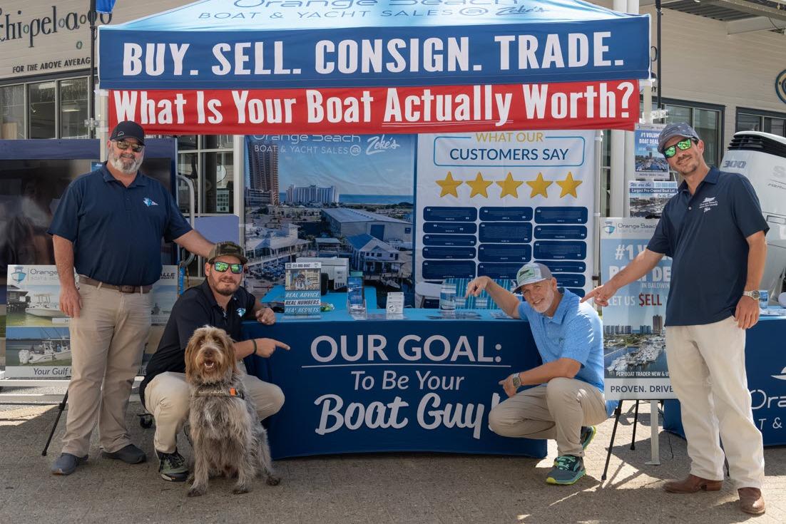 Boat sales team at promotional booth with "Buy. Sell. Consign. Trade." banner.
