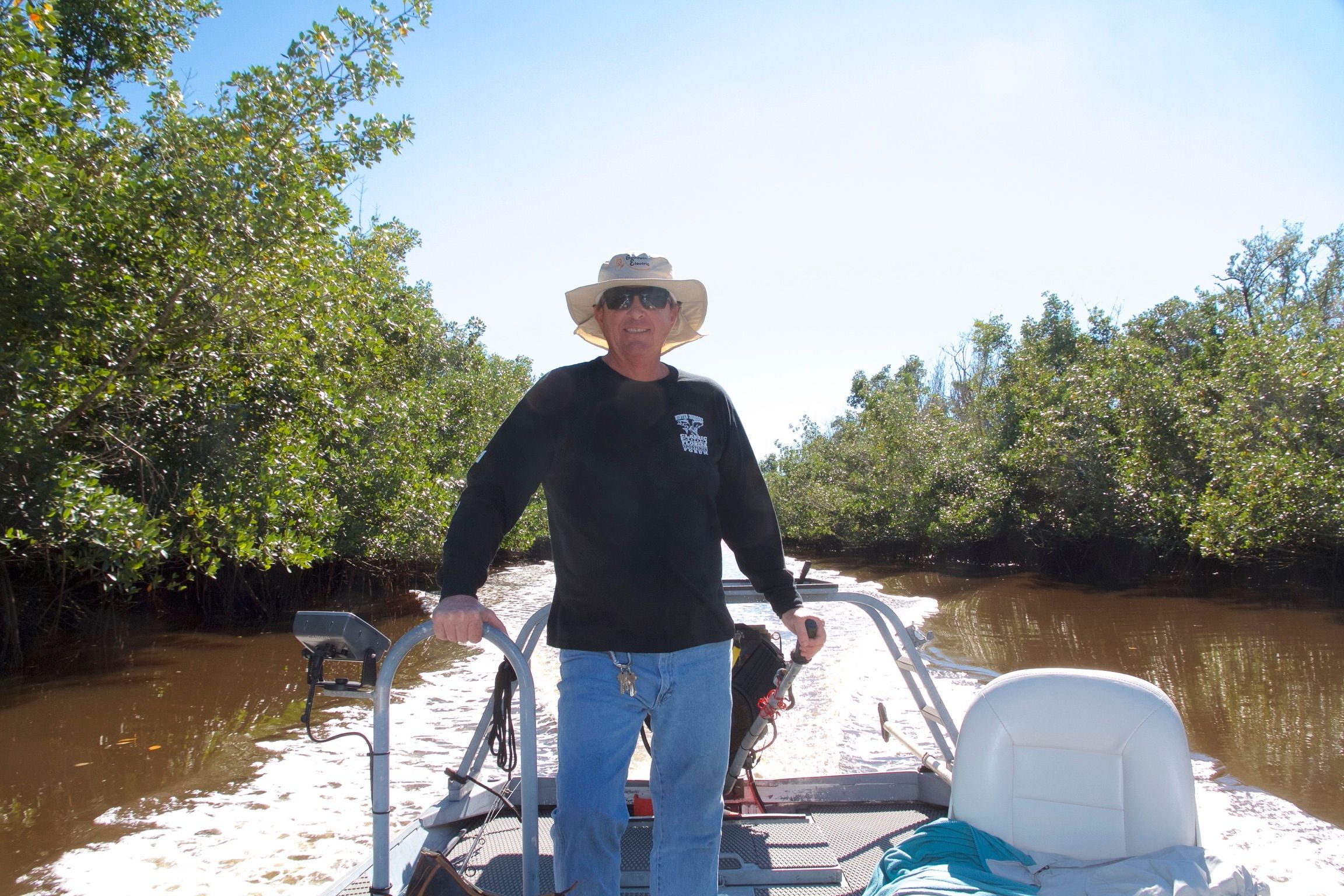 A man steers a Go-Devil 16x56 fishing boat on a narrow river, surrounded by trees.