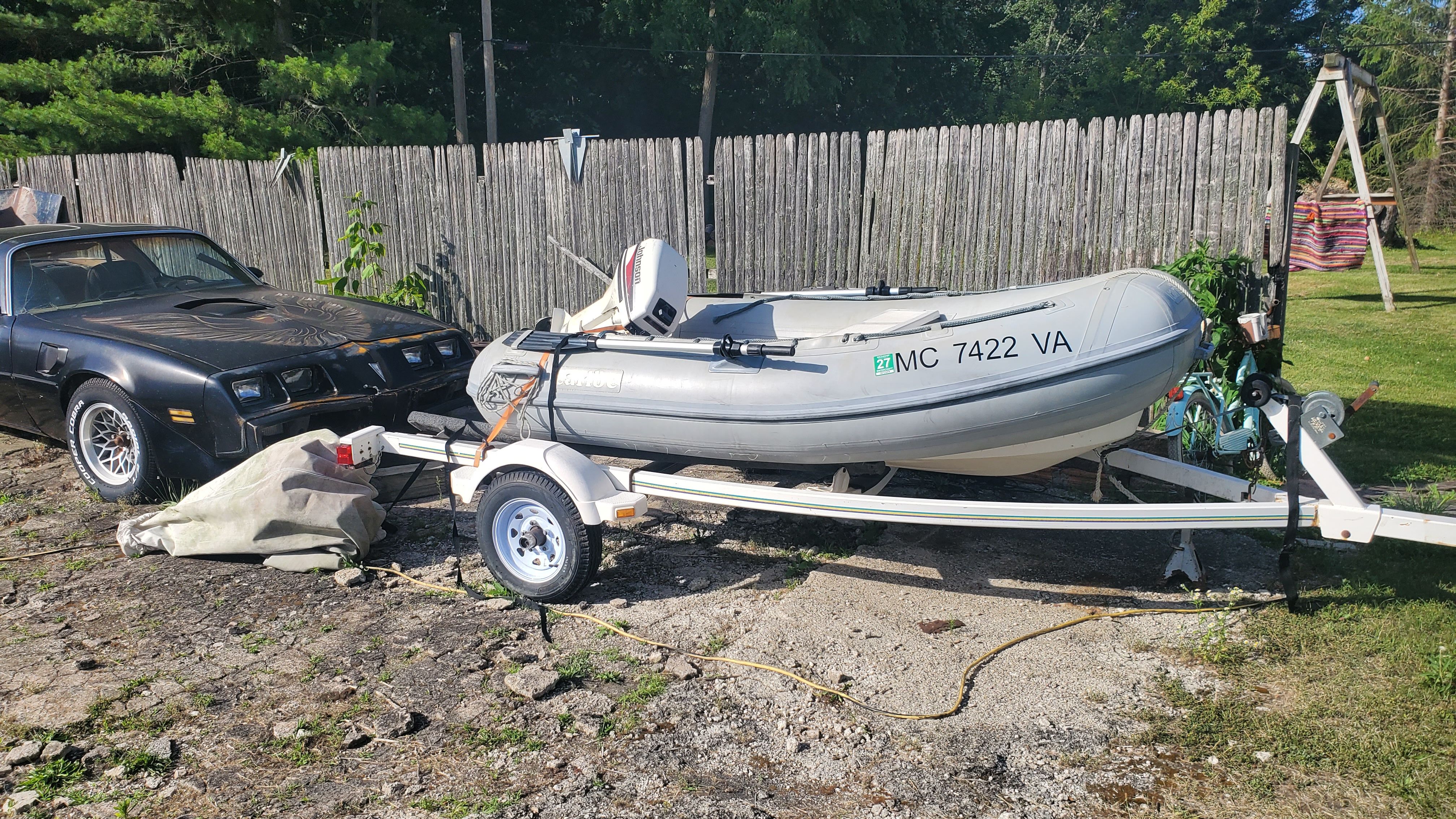 A gray inflatable boat on a trailer next to a black car.