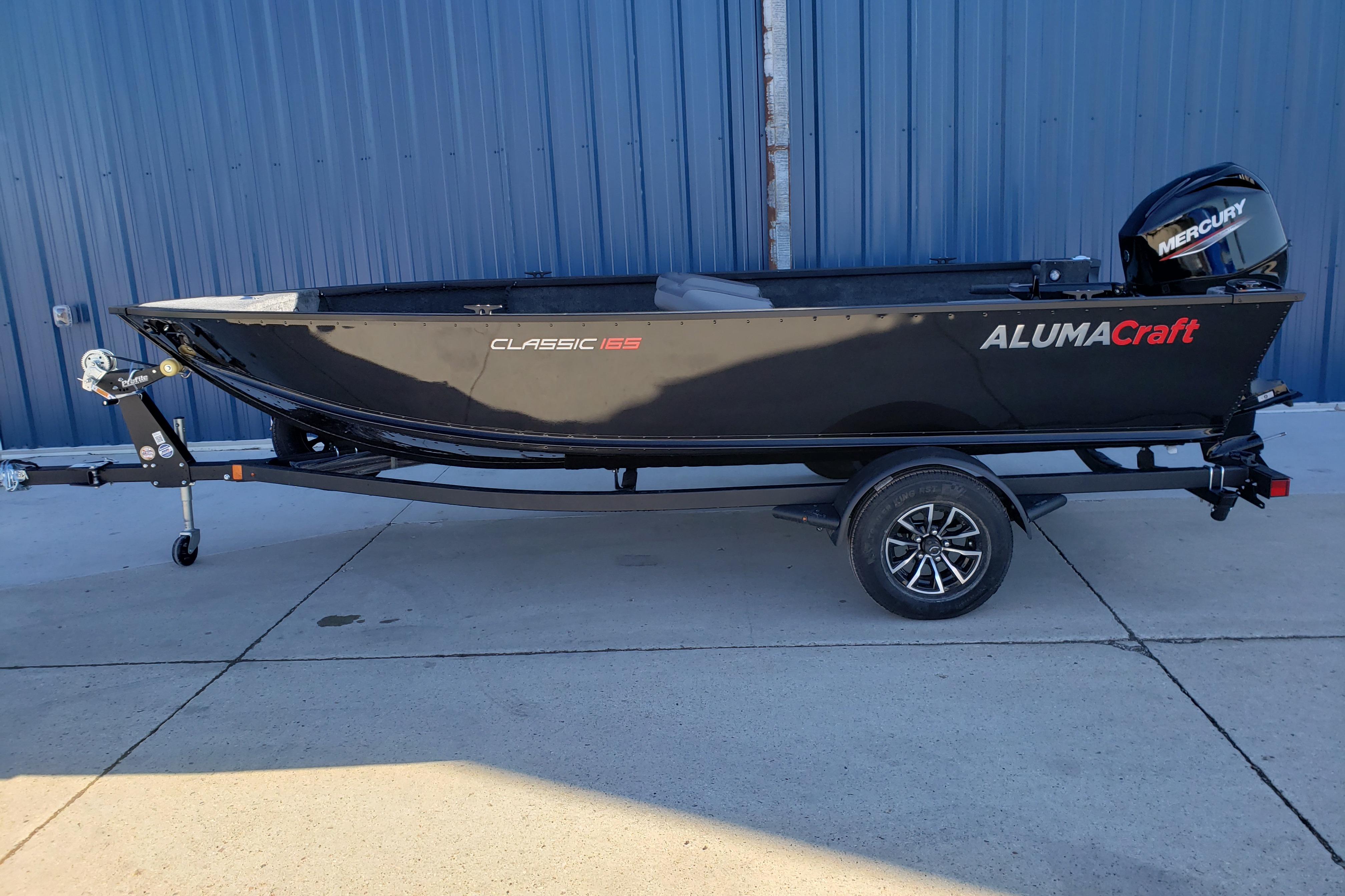 2026 Alumacraft Classic 165 Tiller boat on trailer, parked by a blue wall.