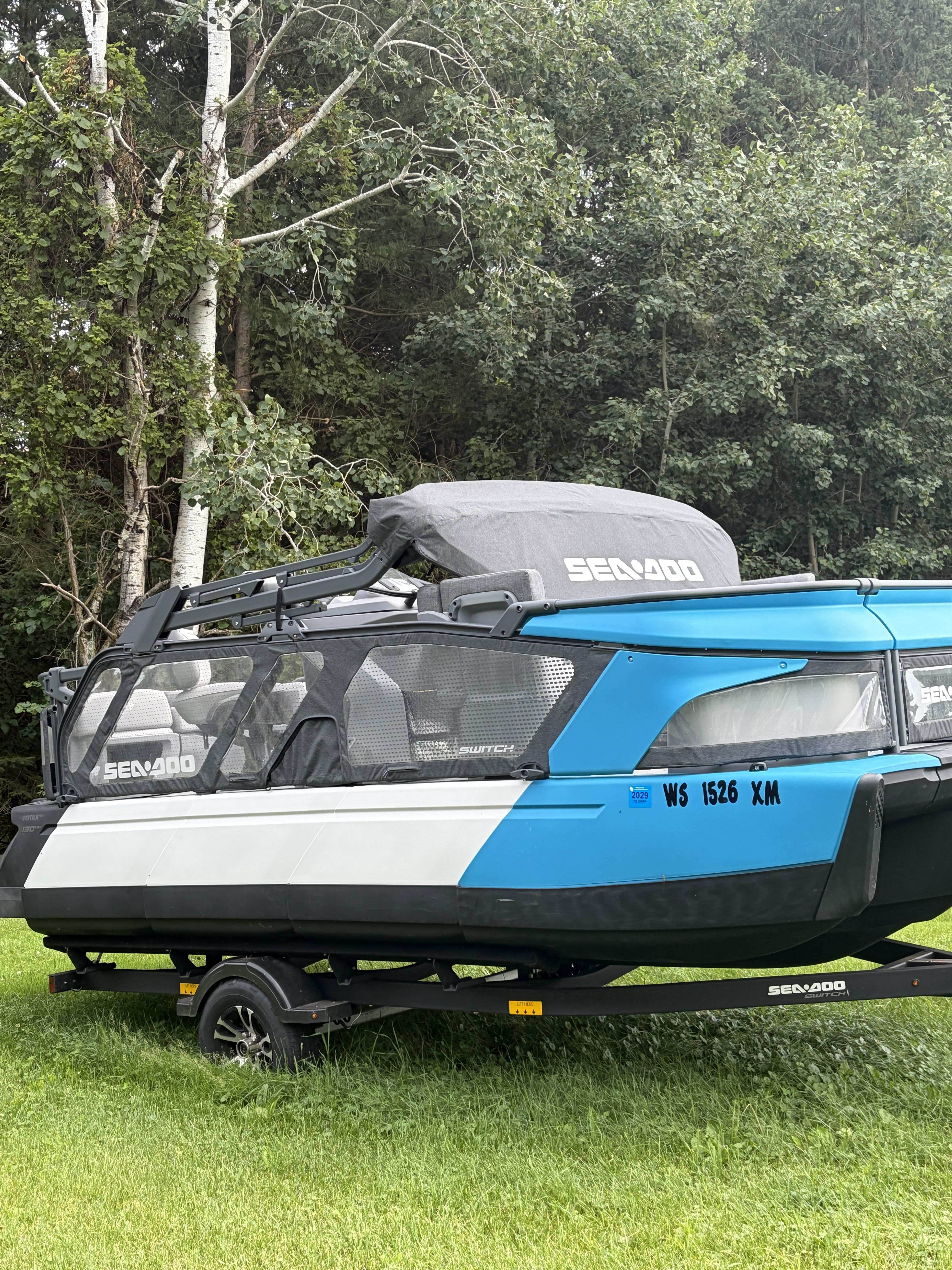 A blue and white Sea Doo Switch boat on a trailer, set on grass with trees behind.