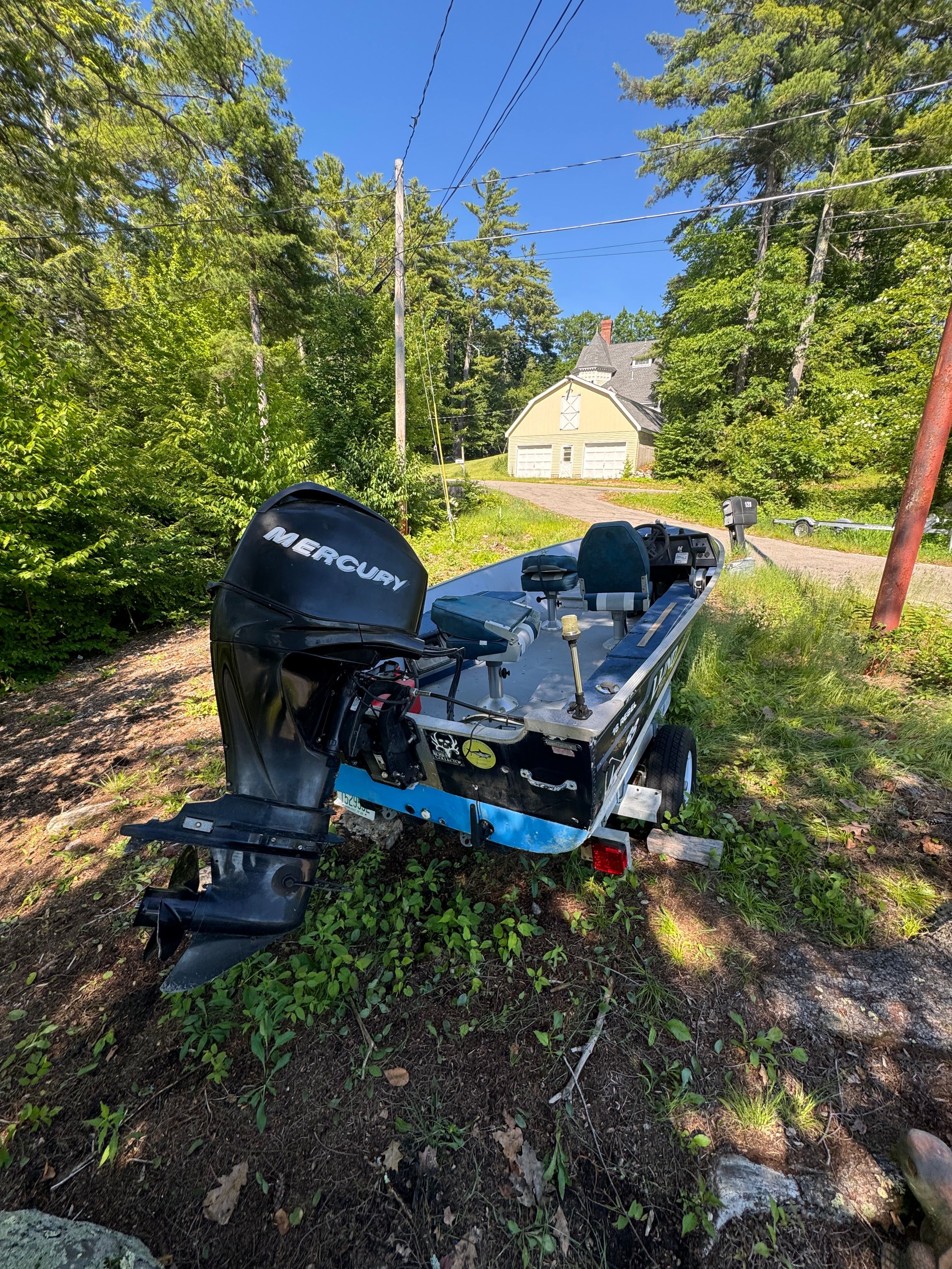 A boat with a Mercury engine is on a trailer in a wooded area.