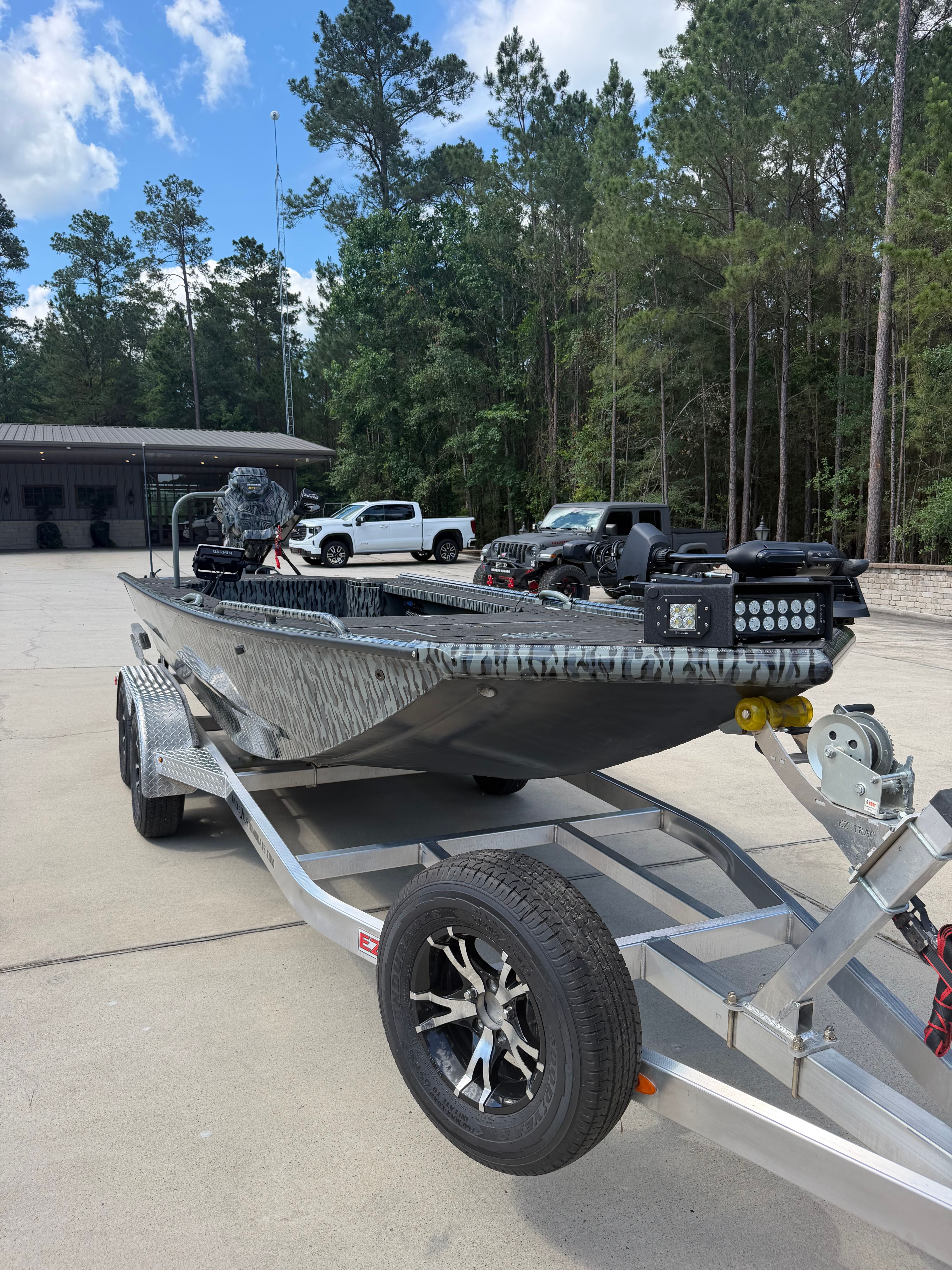 A gray boat on a trailer, surrounded by trees and vehicles.