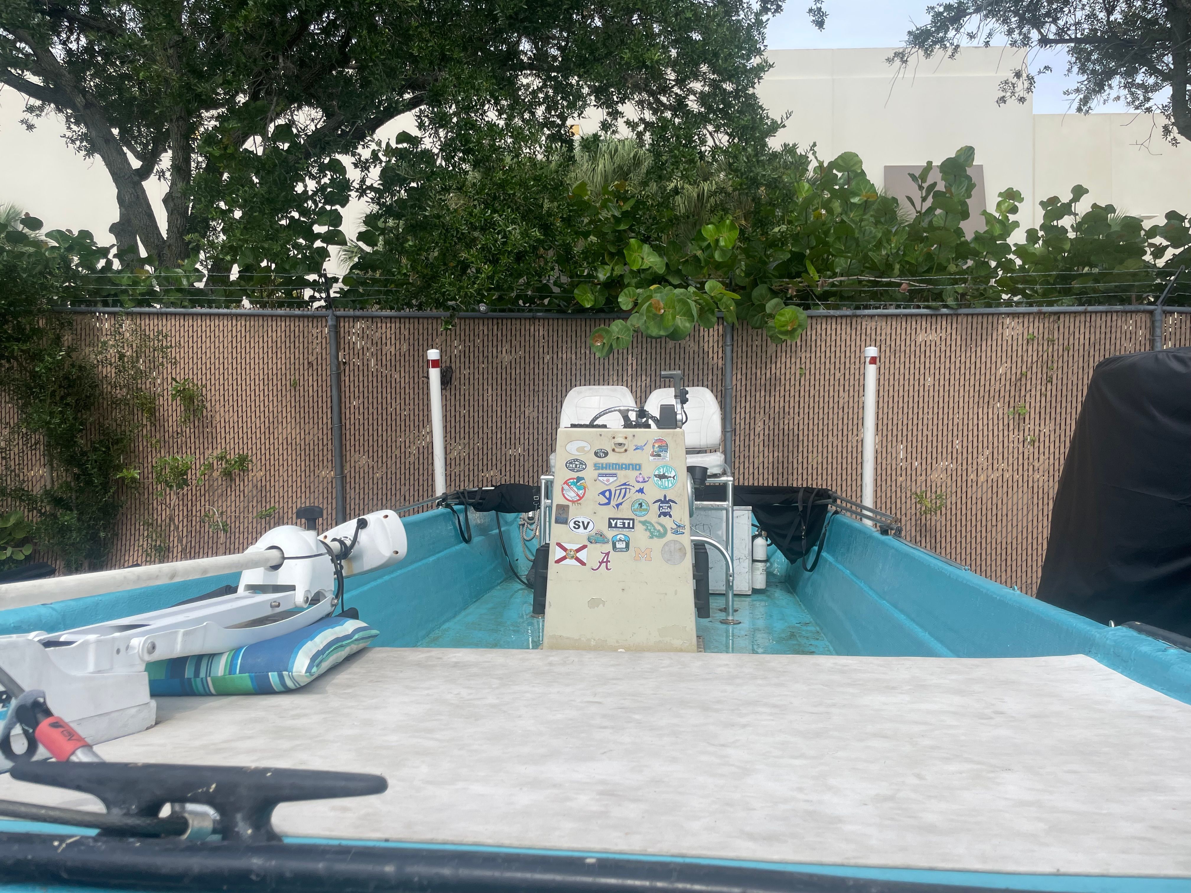 A blue boat with stickers on the console, surrounded by trees and a fence.
