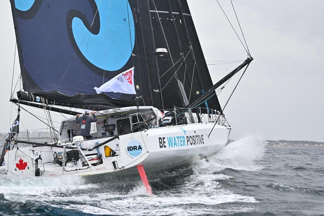 Sailing yacht IMOCA 60 navigating waves, displaying "Be Water Positive" slogan.