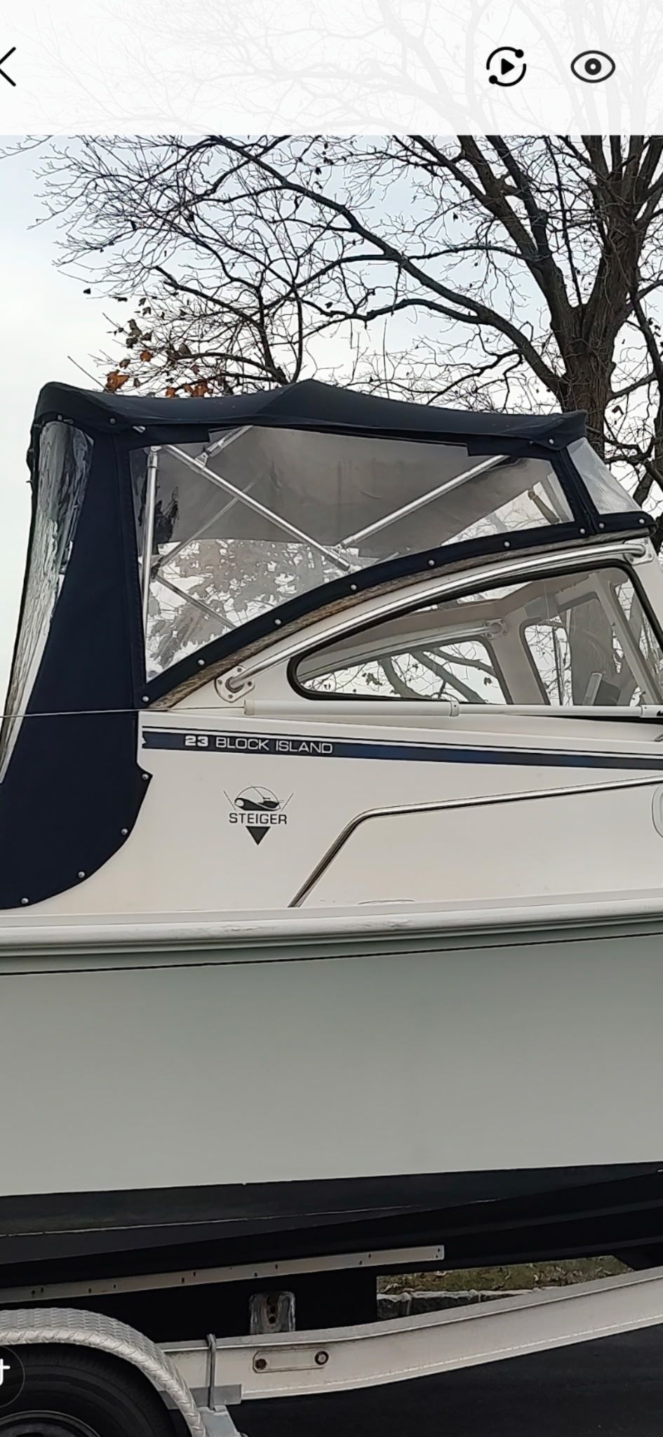 A Steiger Craft boat with a blue cover on a trailer, surrounded by bare trees.
