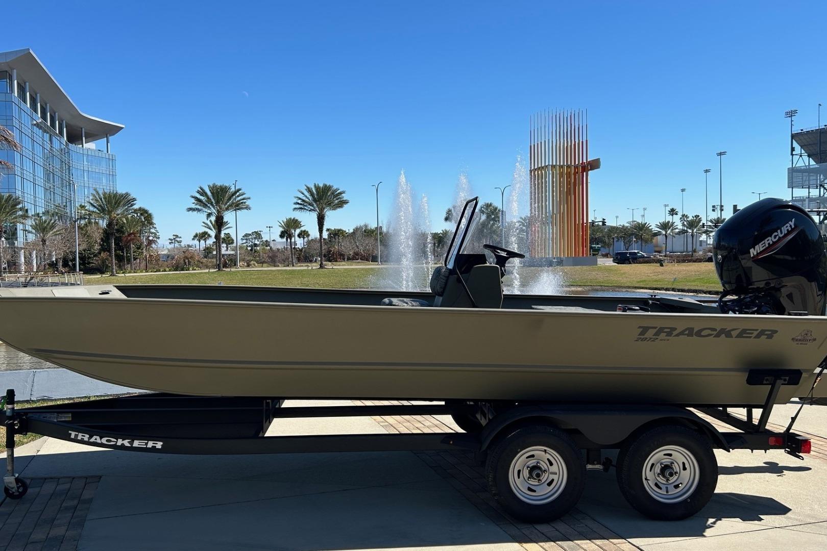 2026 Tracker Grizzly 2072 CC boat on trailer, parked near fountain and modern buildings.