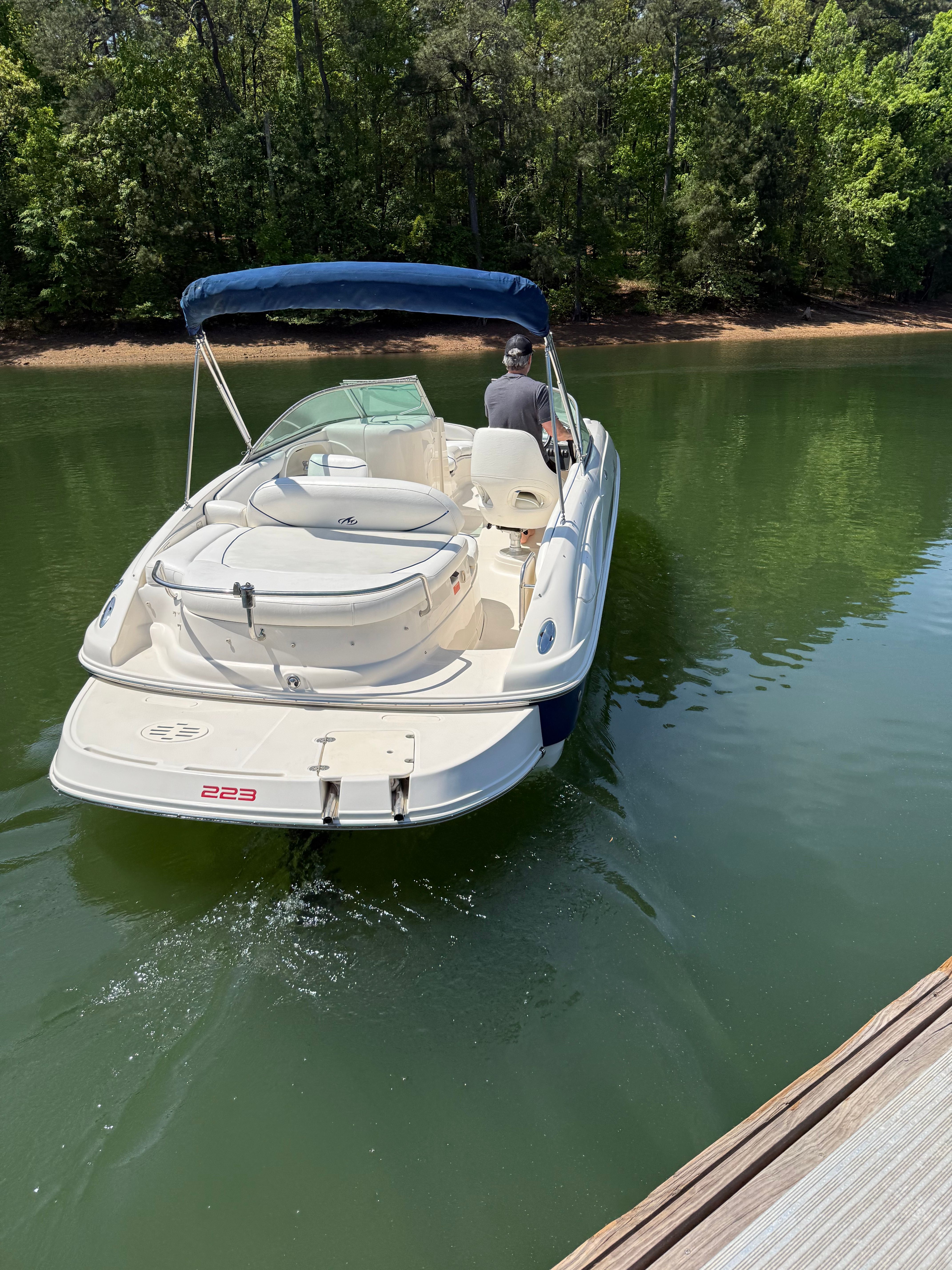 A white boat with a blue canopy navigates a calm lake, surrounded by trees.