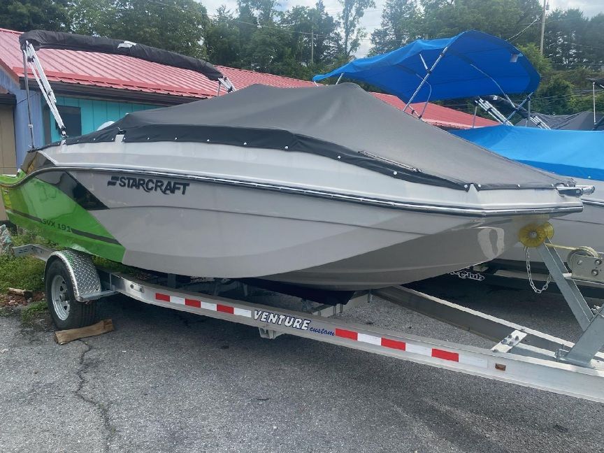 Starcraft SVX 191 OB 2023 on a trailer with a gray cover, parked outdoors.