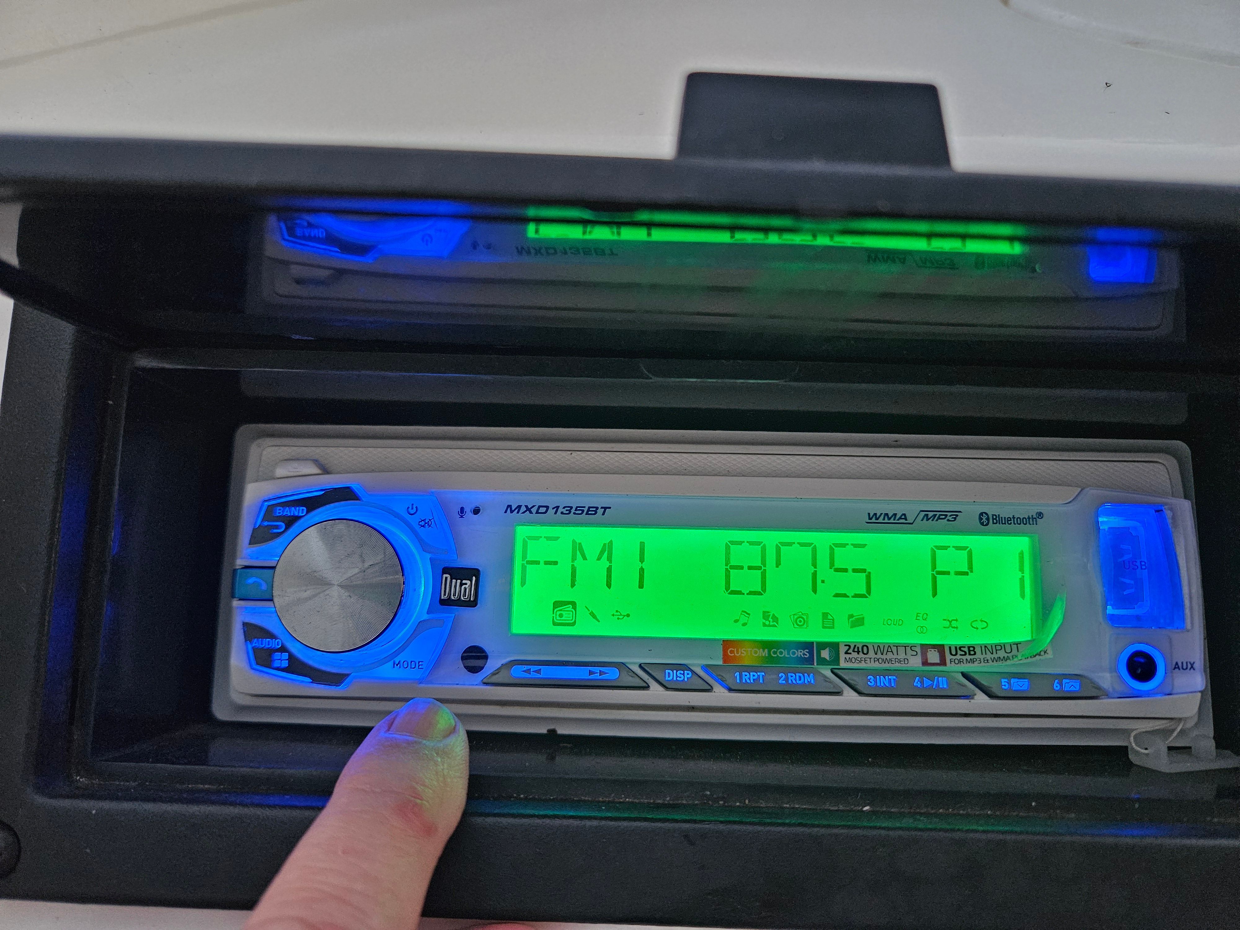 A modern car stereo with a glowing display shows FM 87.5.