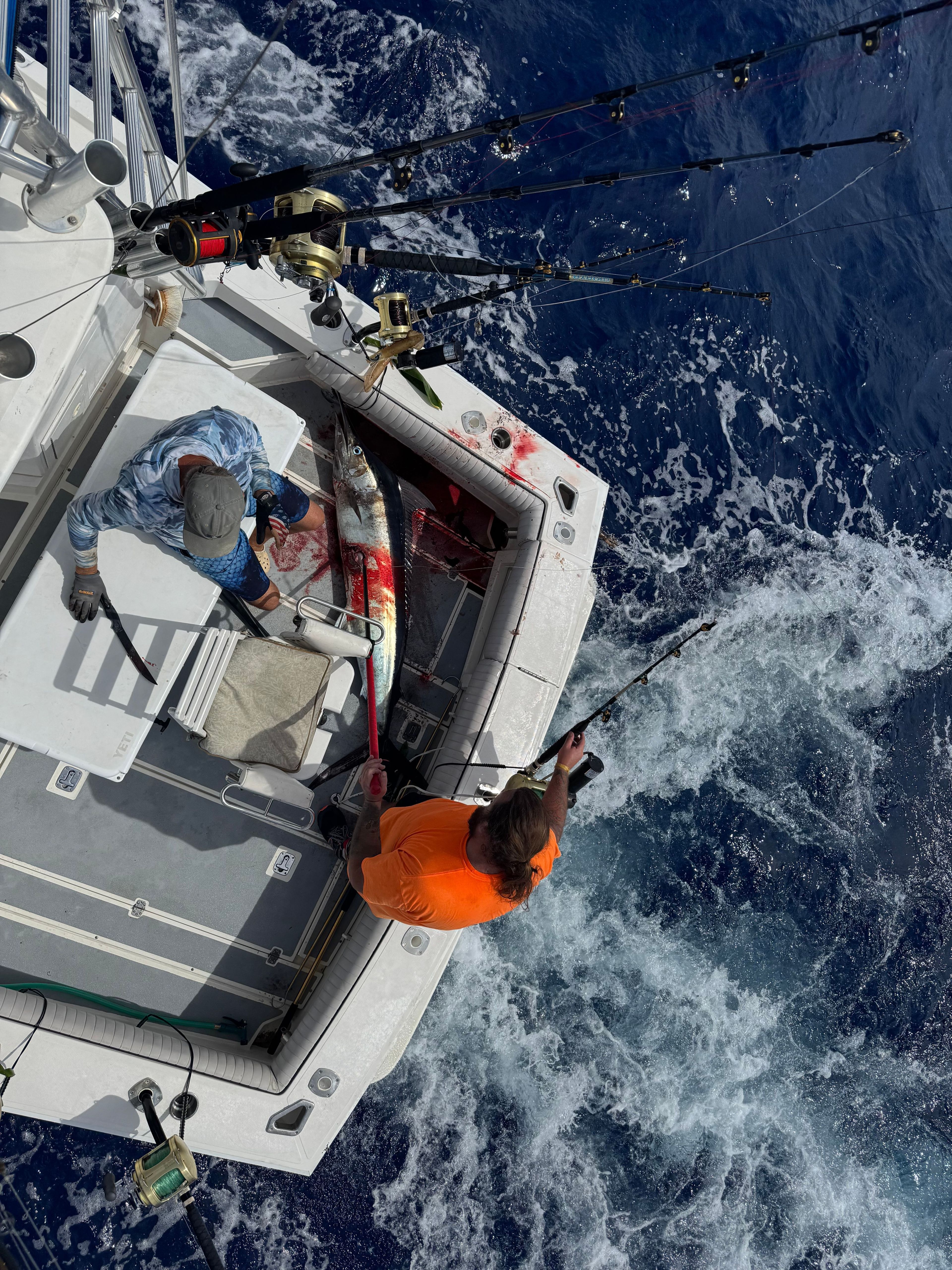 Two people are fishing on a boat in open water. A large fish lies on the deck.