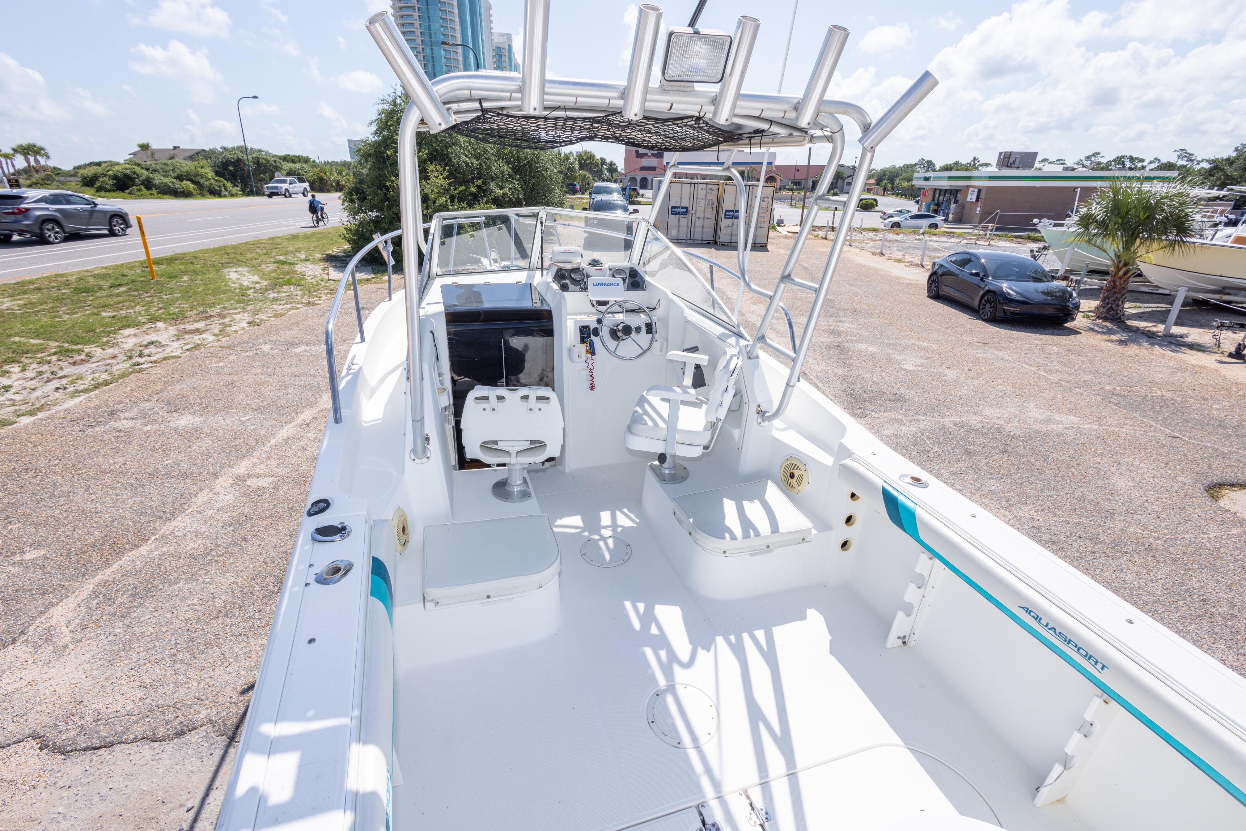 1999 Aquasport 225 Explorer boat with open deck, steering console, and seating, parked near a road.