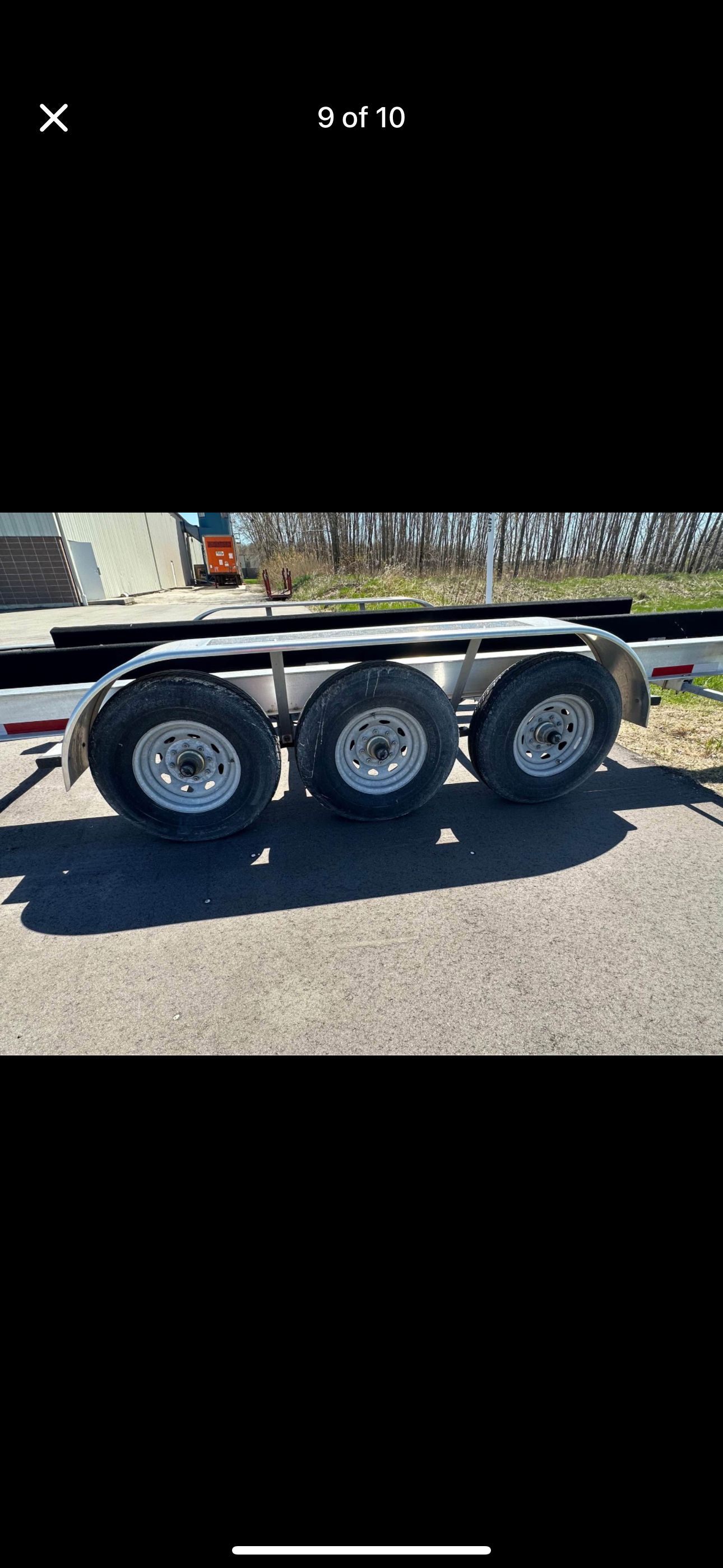 A triple-axle boat trailer is parked on a paved area, surrounded by trees.