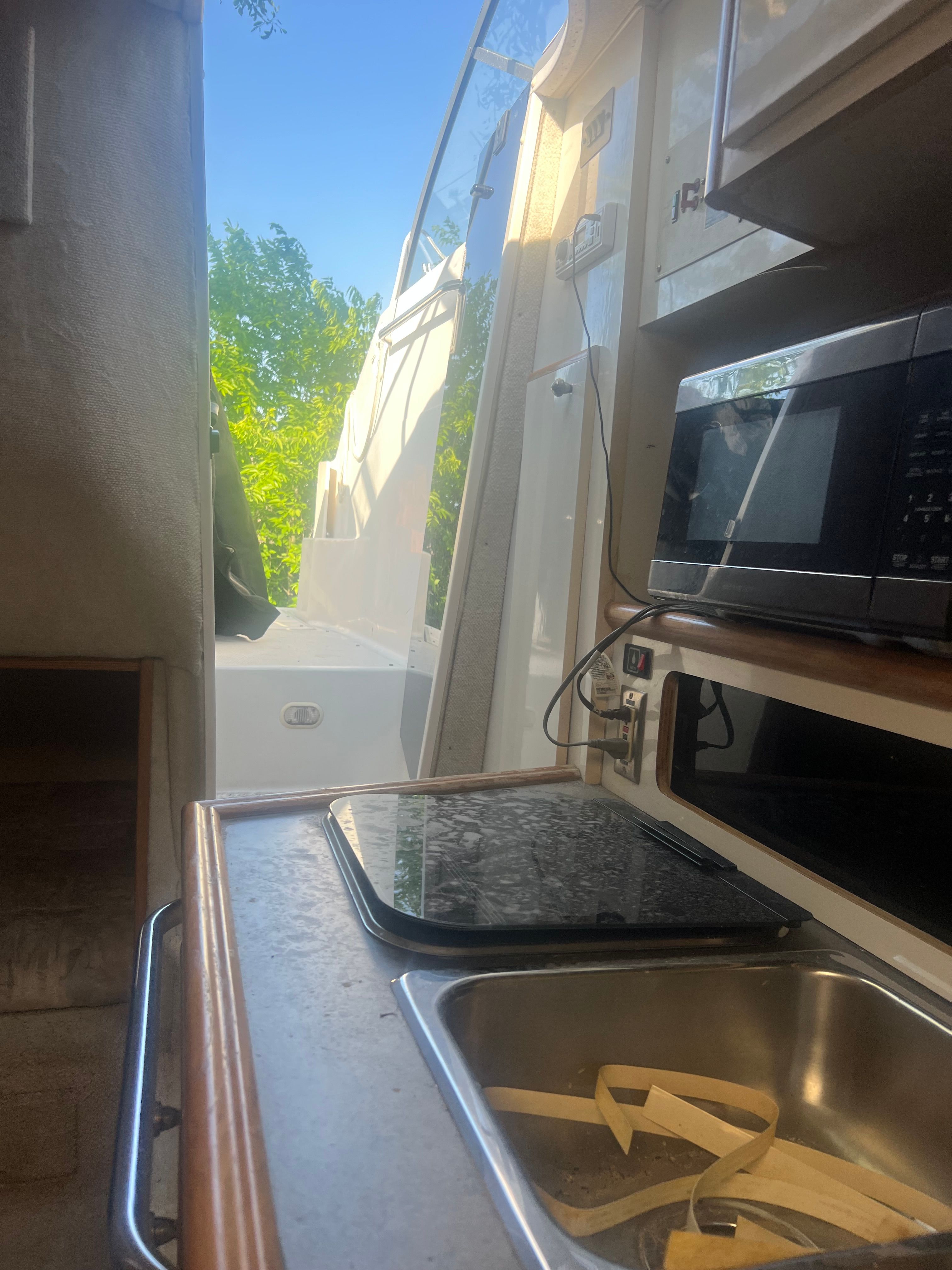 Interior view of a boat with a sink, stove, and microwave. Trees are visible outside.