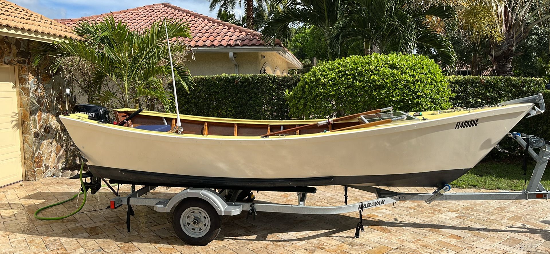 A cream-colored dory boat on a trailer, surrounded by palm trees and a house in the background.