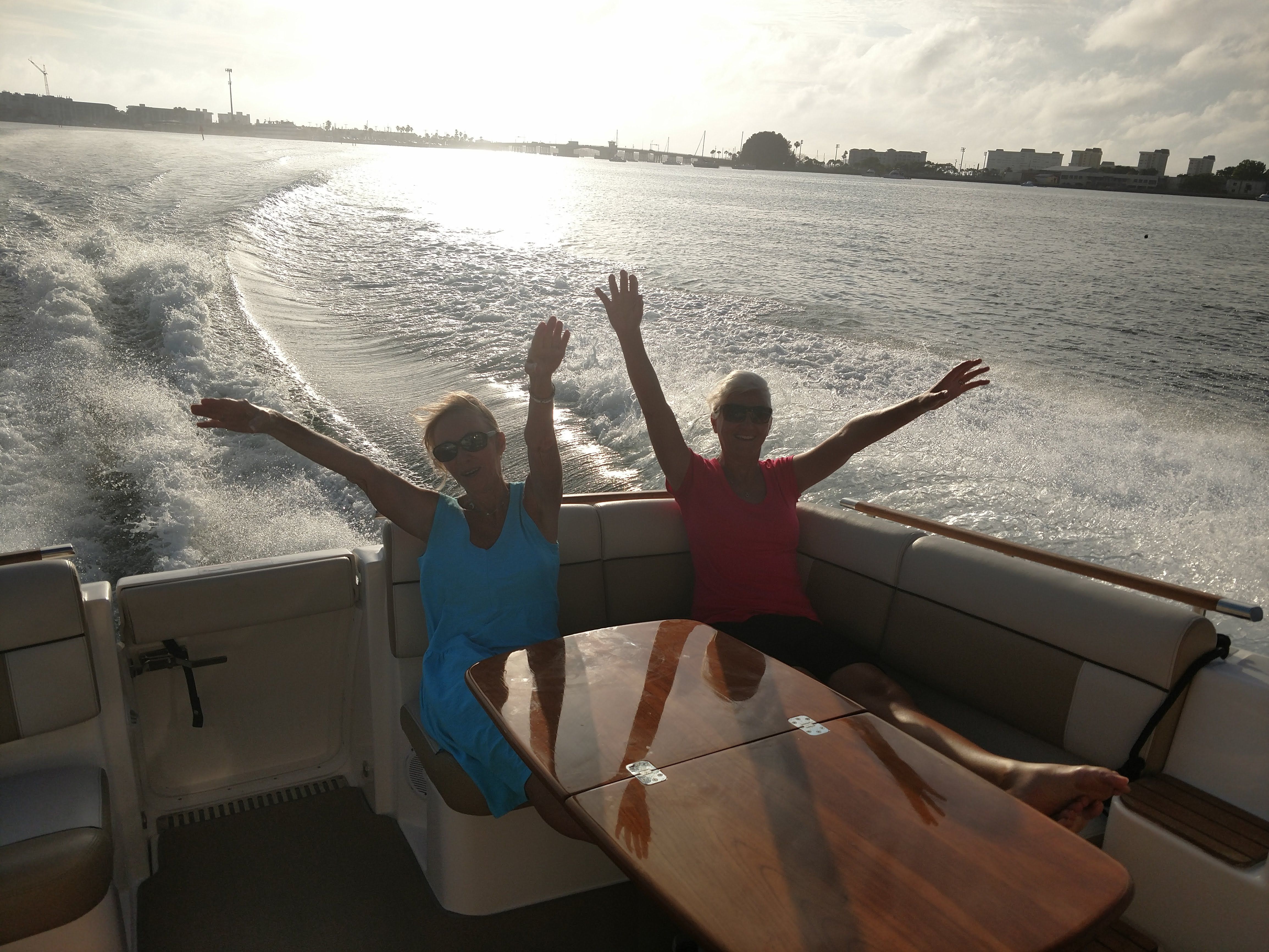Two people sit on a boat speeding over water, arms raised in excitement.