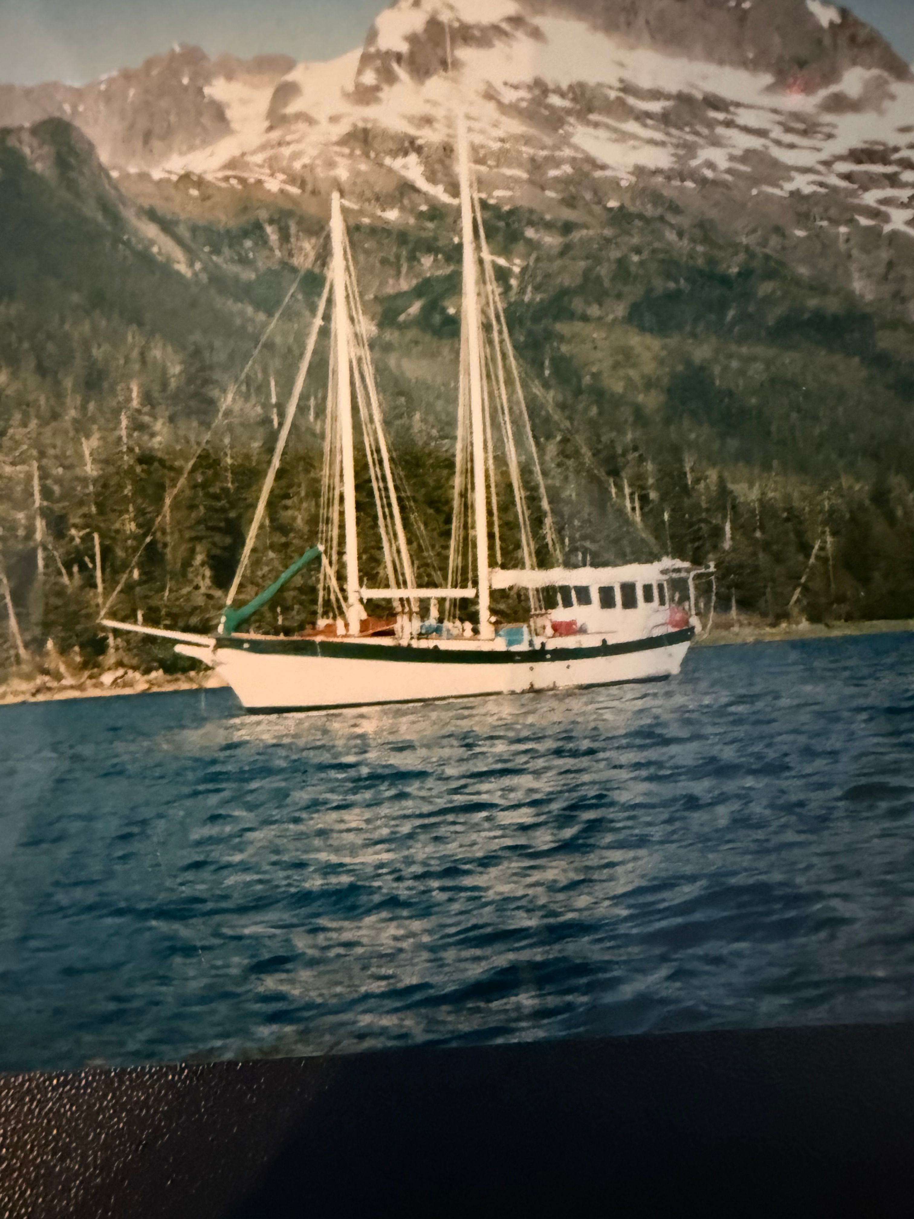 A white schooner sails near a forested coastline with snow-capped mountains in the background.