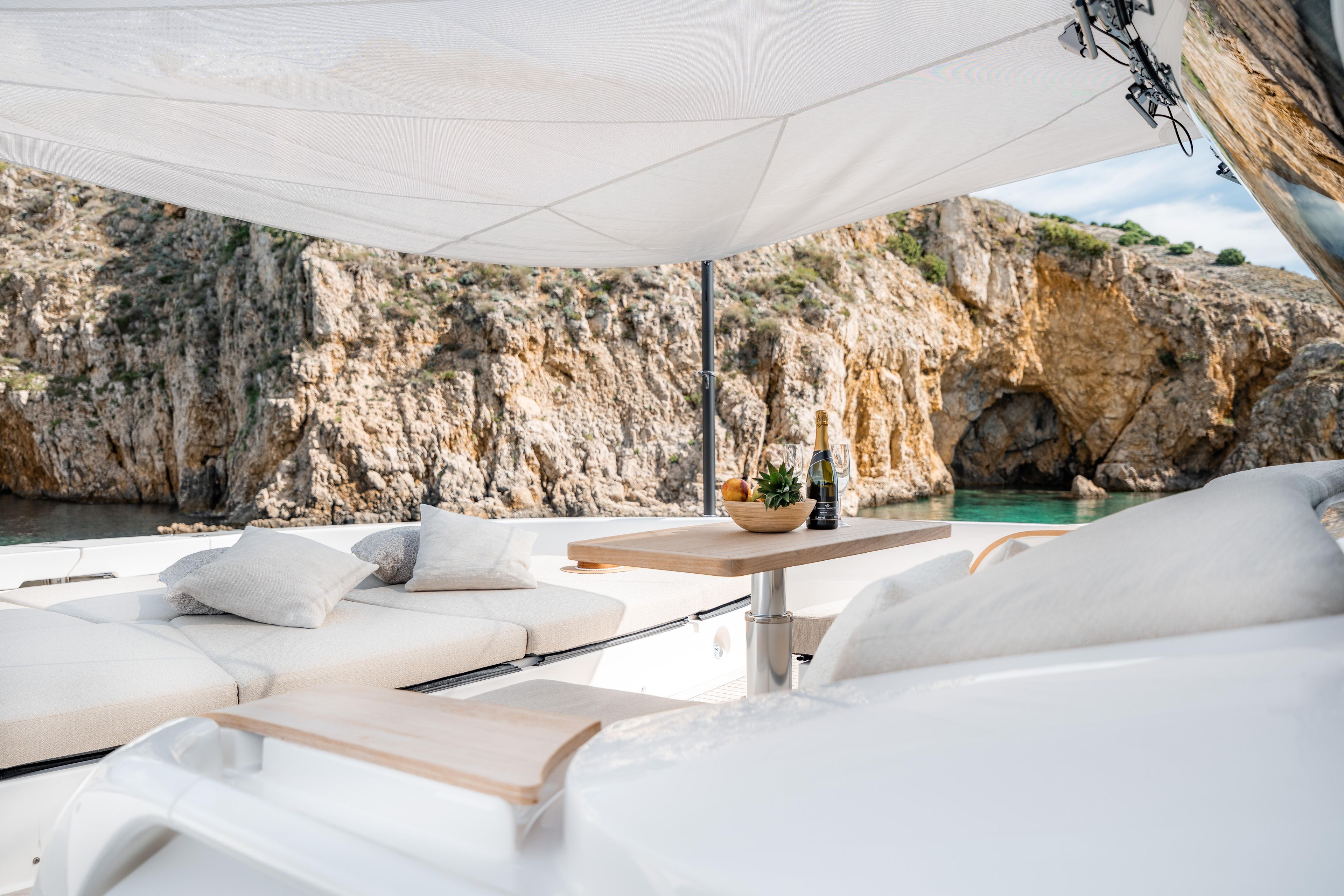 Luxurious Bluegame BG74 yacht deck with cushions, table, and scenic rocky coastline view.