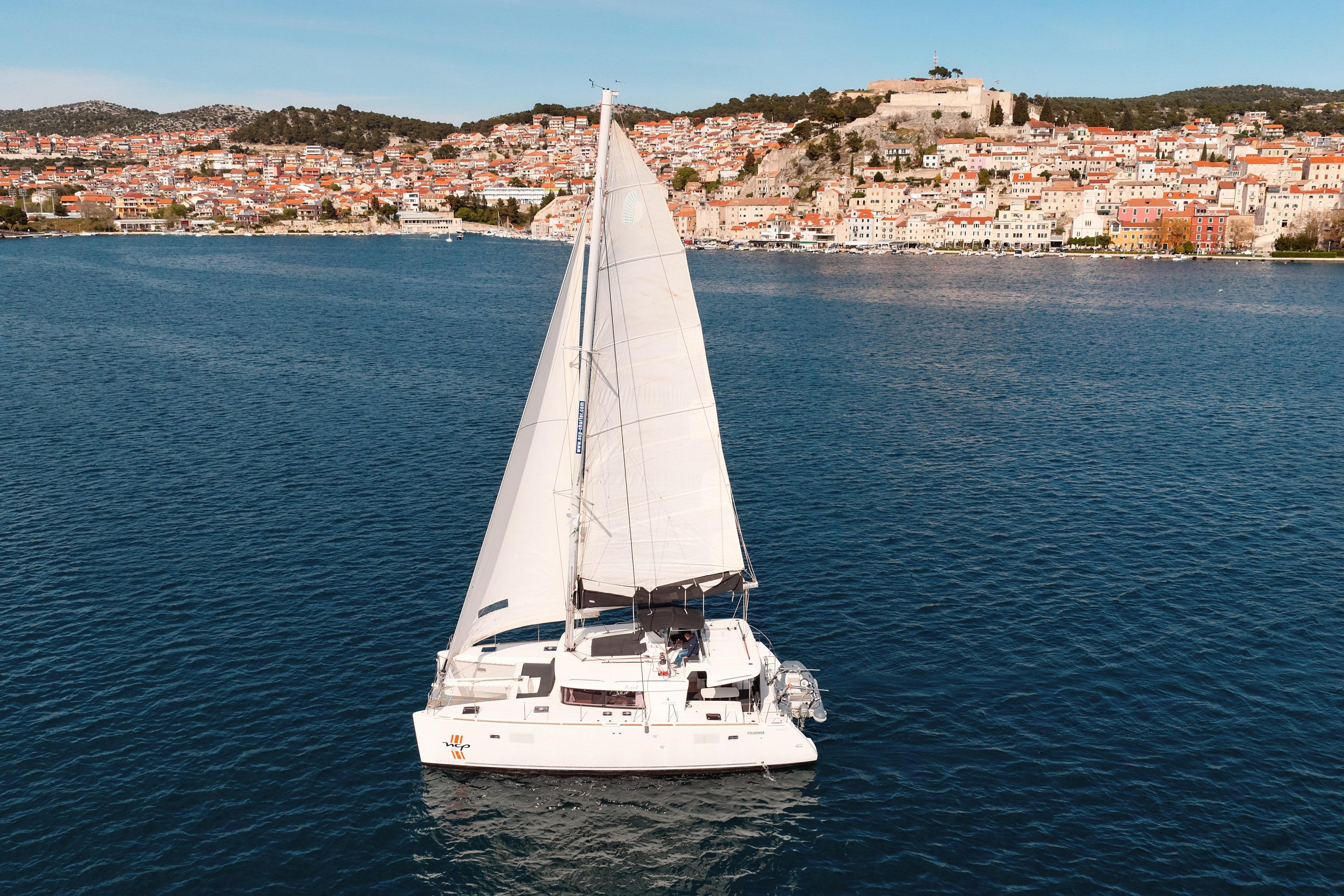 2018 Lagoon 450 sailing in coastal waters with scenic town backdrop.