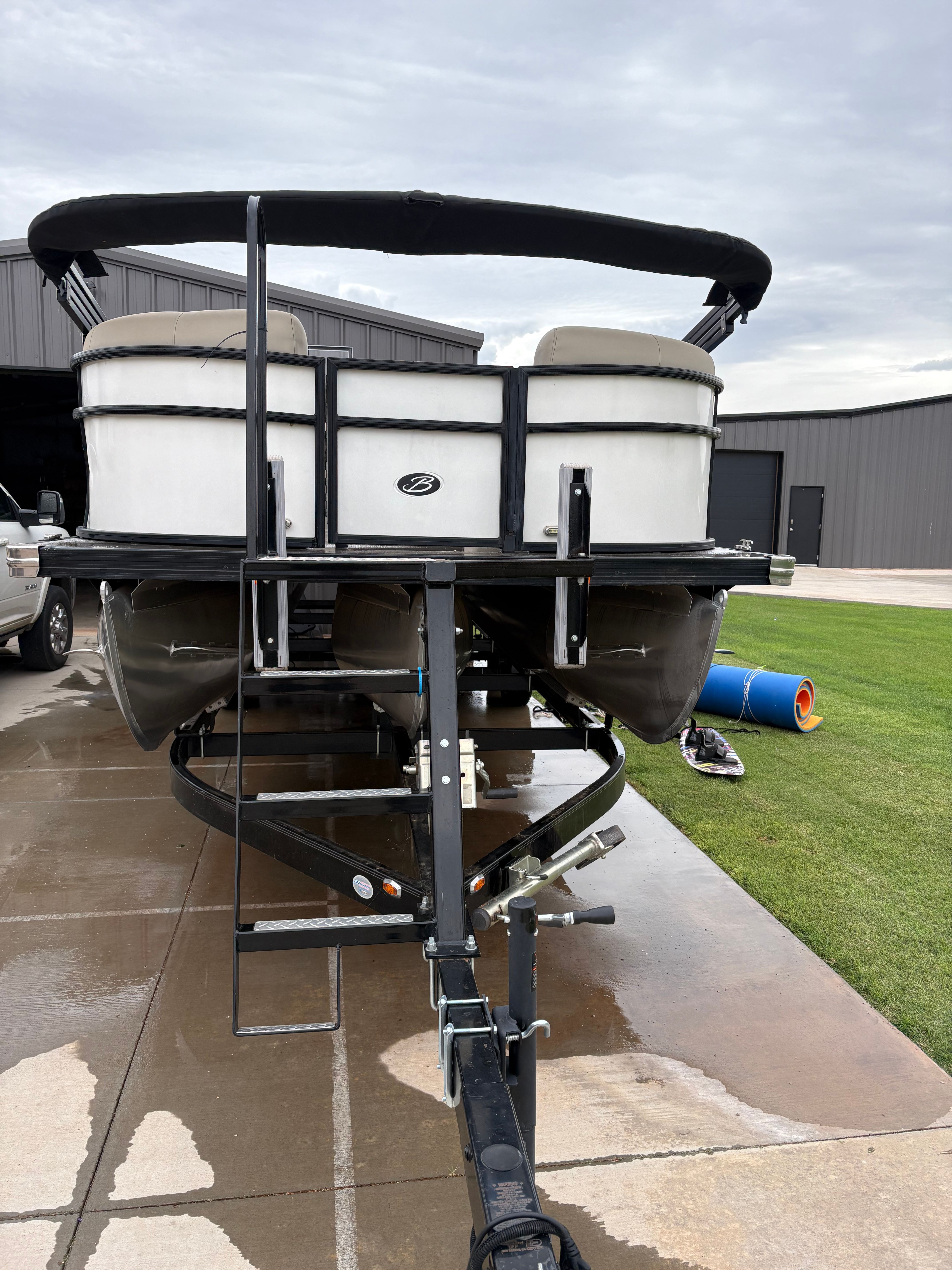 A Barletta pontoon boat on a trailer, parked on a driveway next to a building.