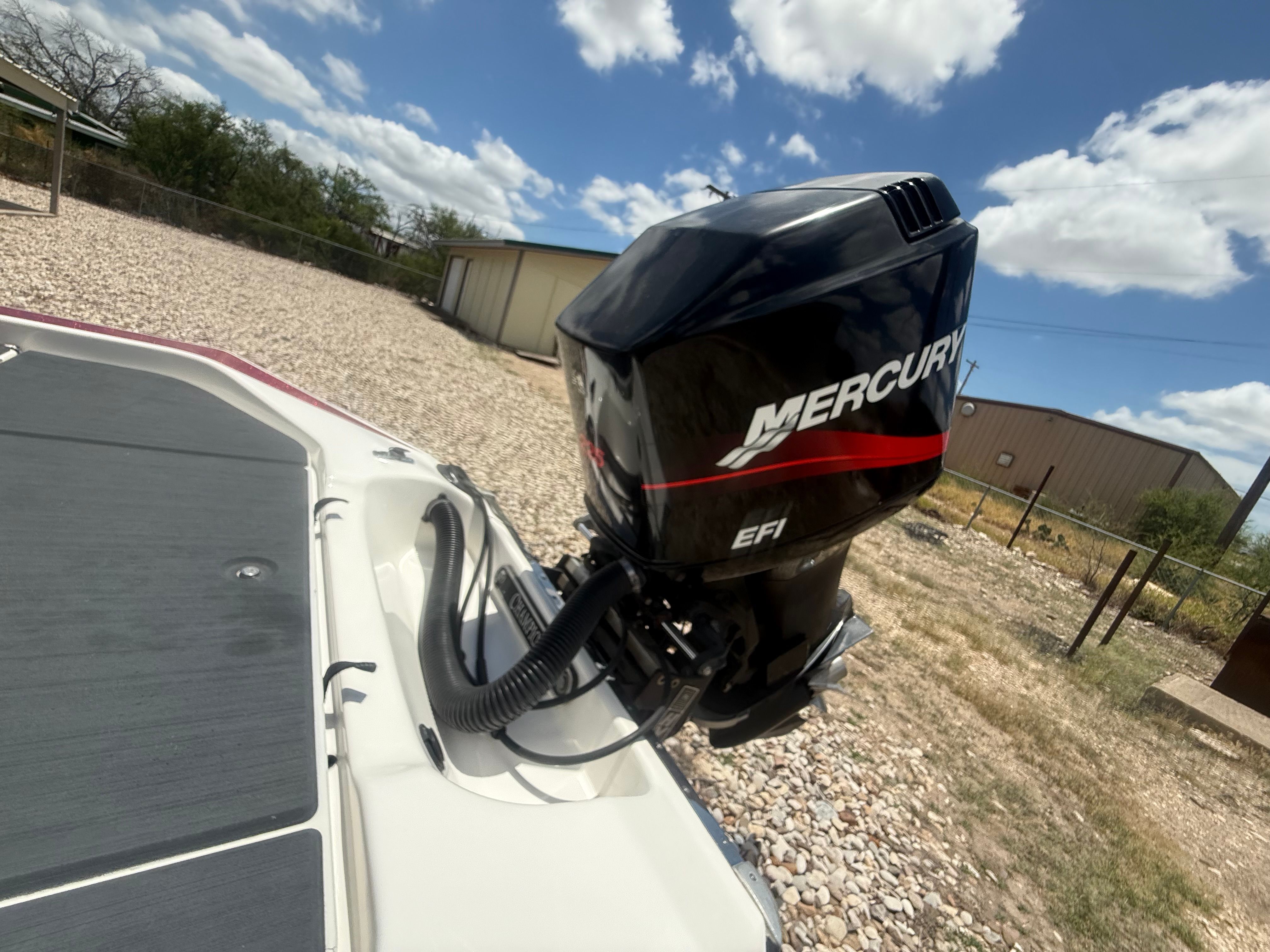 A Mercury EFI outboard motor is mounted on a white boat situated on a gravel lot.