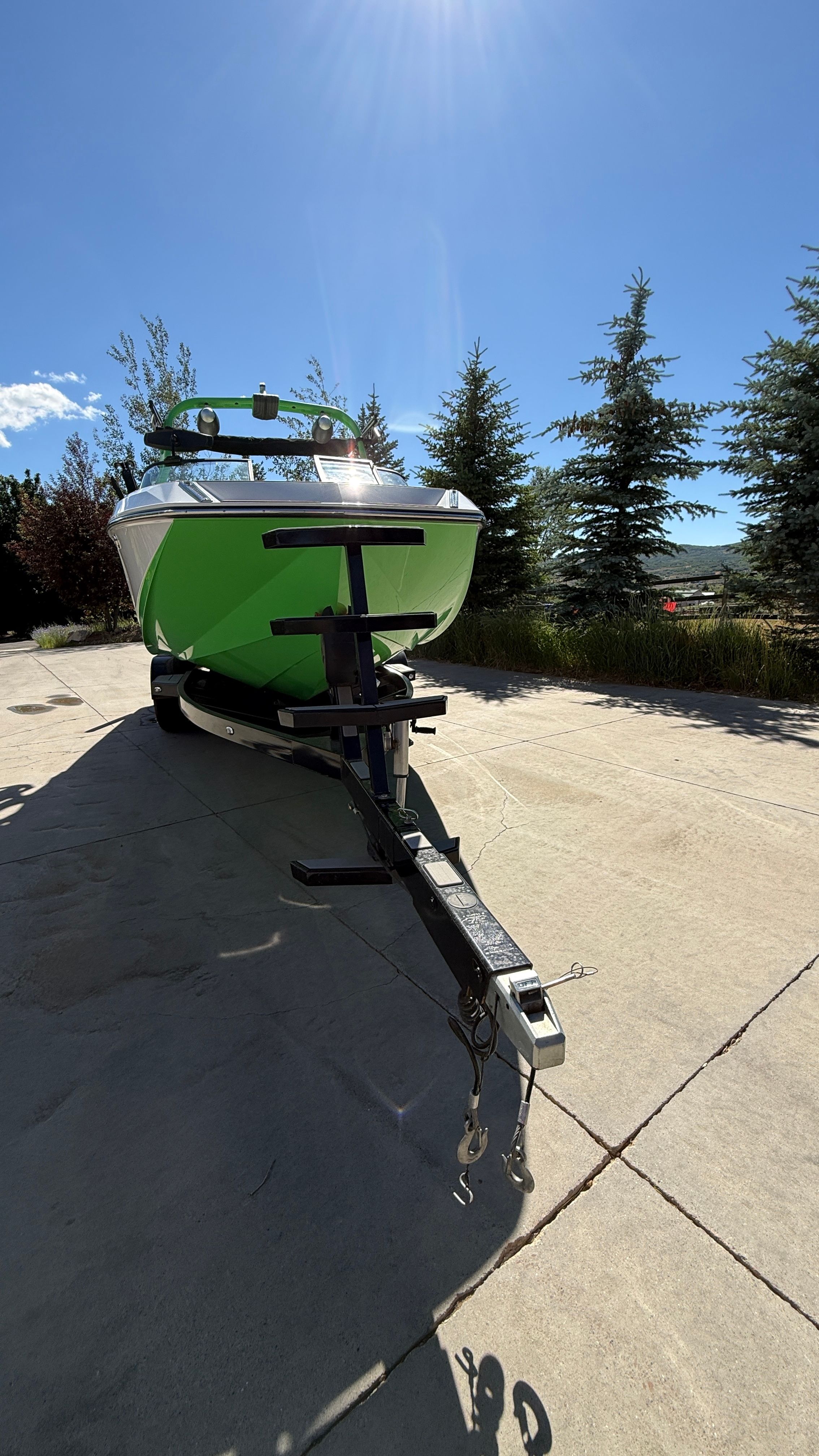 A green boat on a trailer on a paved path, surrounded by trees.