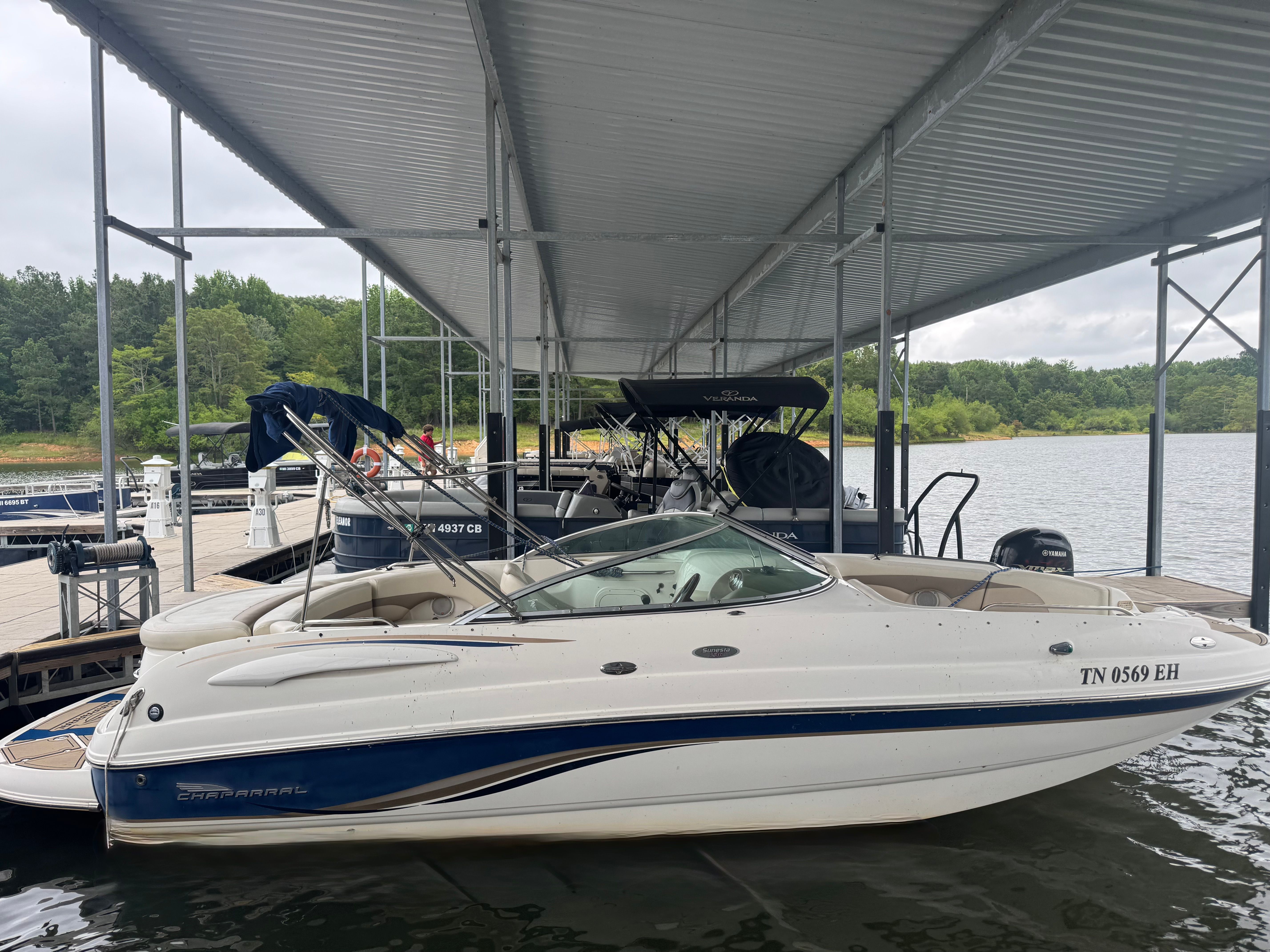A white Chaparral Sunesta 223 boat is docked under a covered pier on a calm lake.