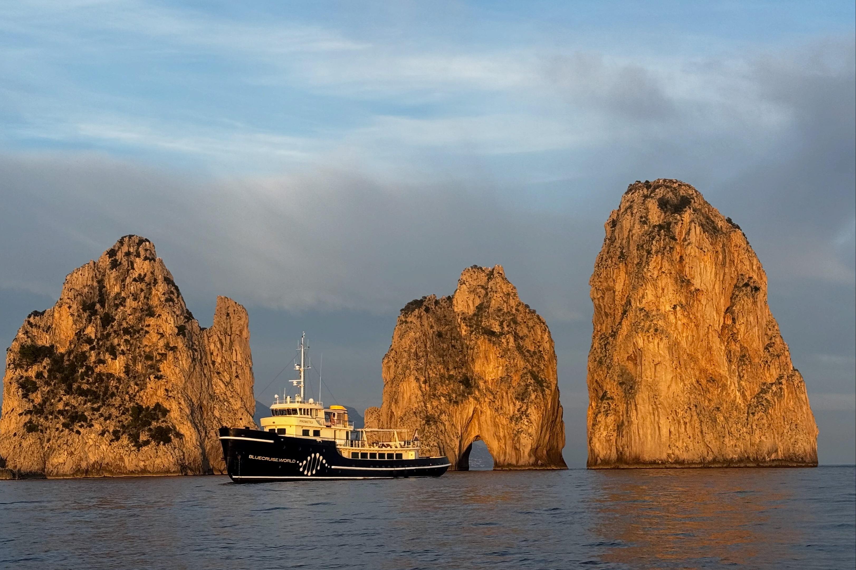 Custom 1956 converted tugboat near towering rock formations at sea during sunset.