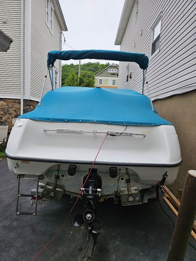 A 1998 Crownline 210 CCR boat with a blue cover, parked between two houses.
